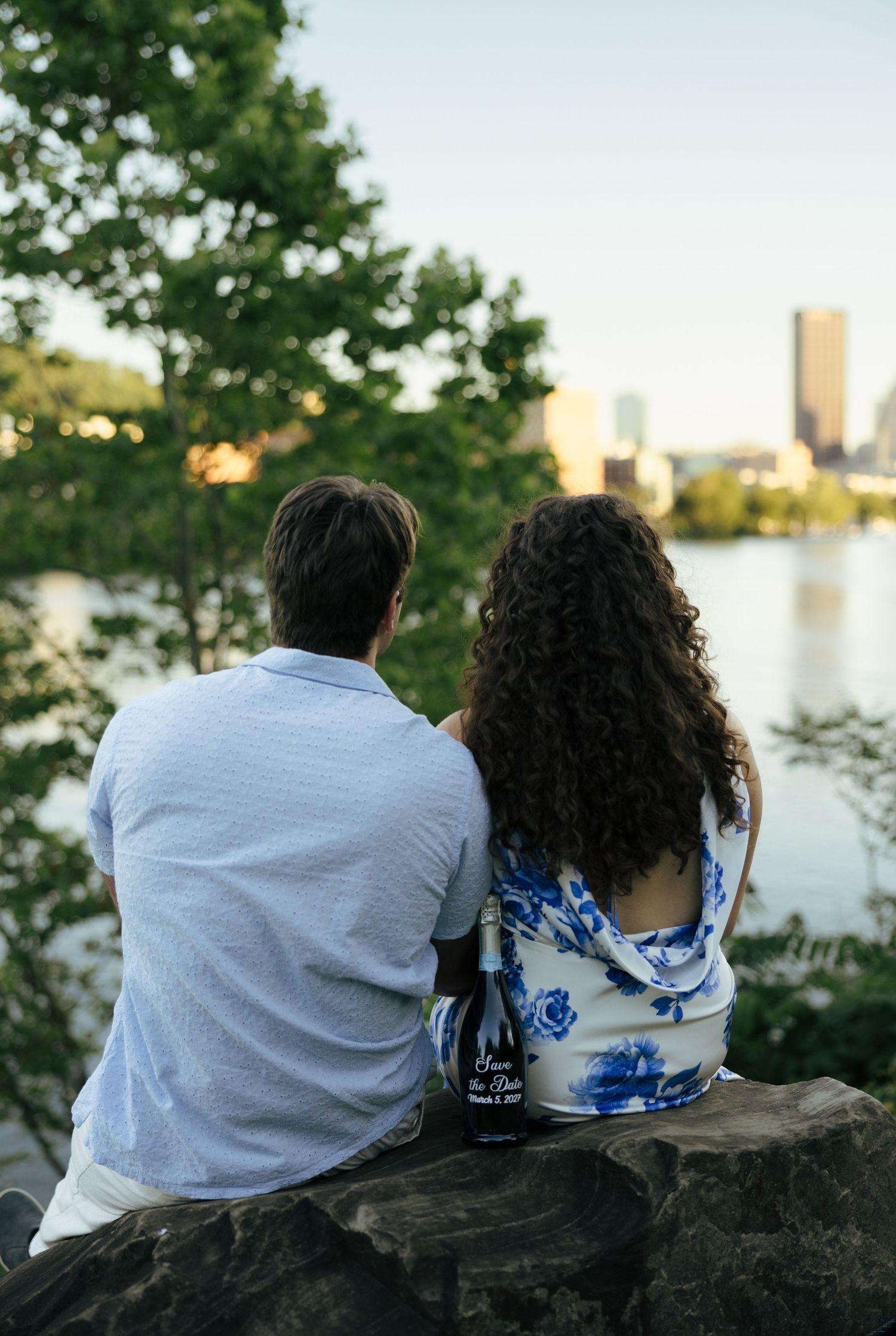 Couple sitting on rocks from behind, admiring city skyline across river holding Save the Date bottle.