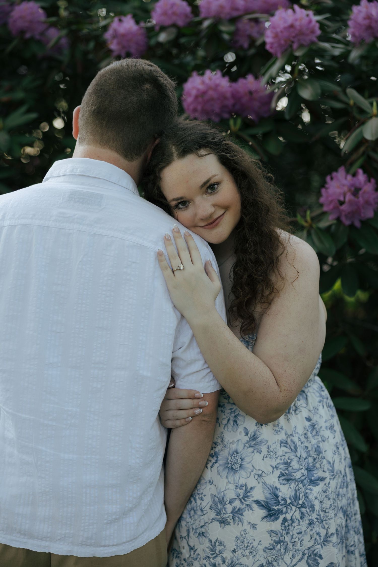 Engaged woman showing ring while embracing partner near blooming pink rhododendron bushes.