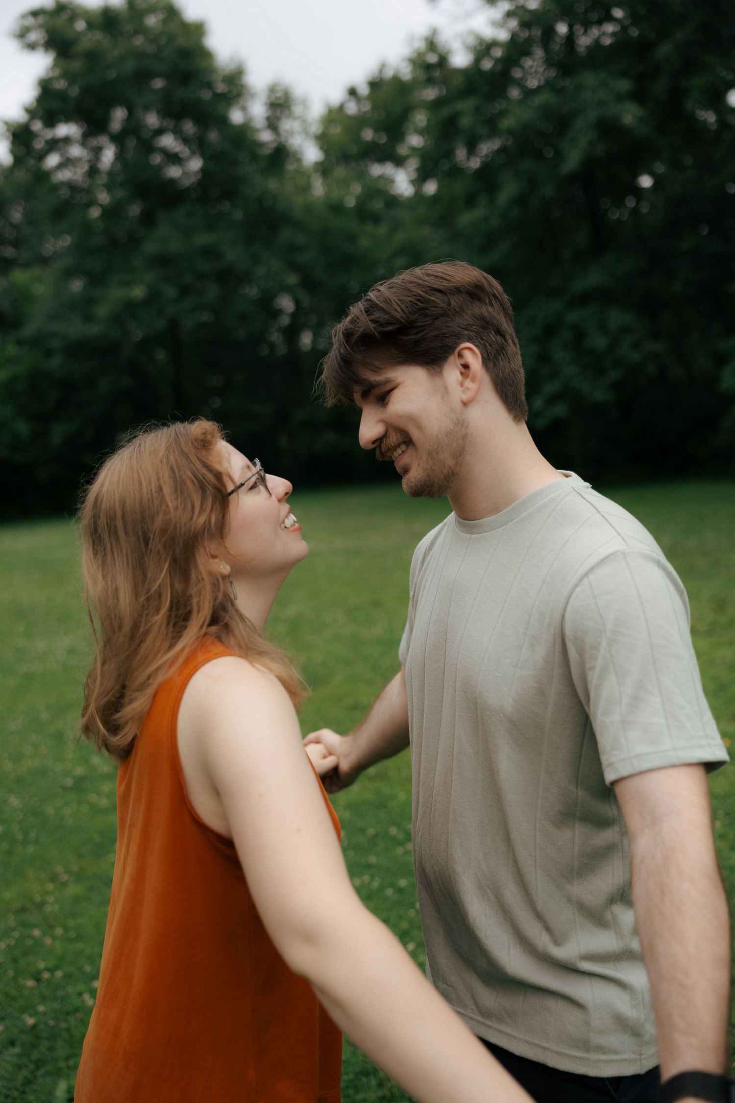 A laughing couple faces each other joyfully while standing together on a green grassy lawn in a park.