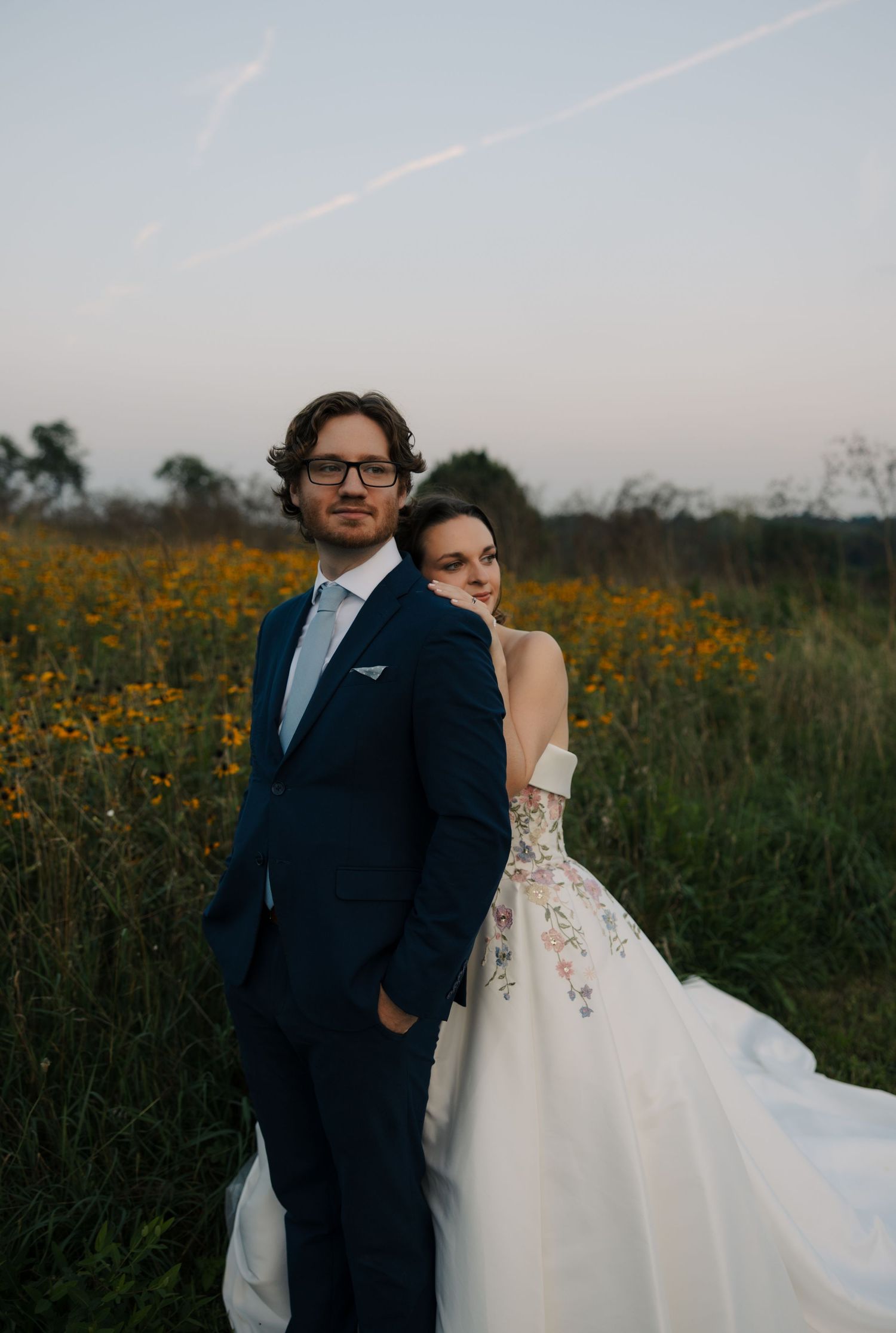 Groom in navy suit and glasses stands with bride leaning on him in floral gown in a flower field.