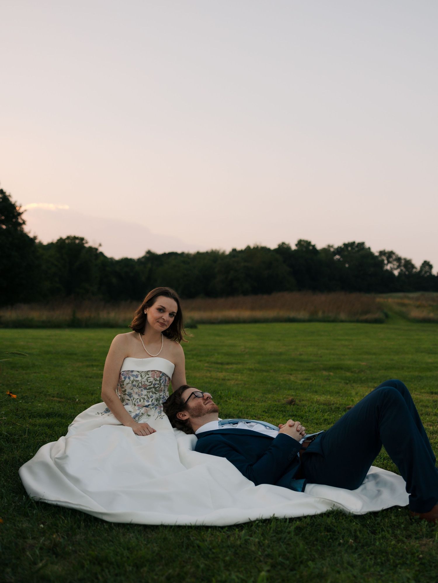 Bride in strapless gown sits on grass as groom in navy suit reclines across her lap at sunset.