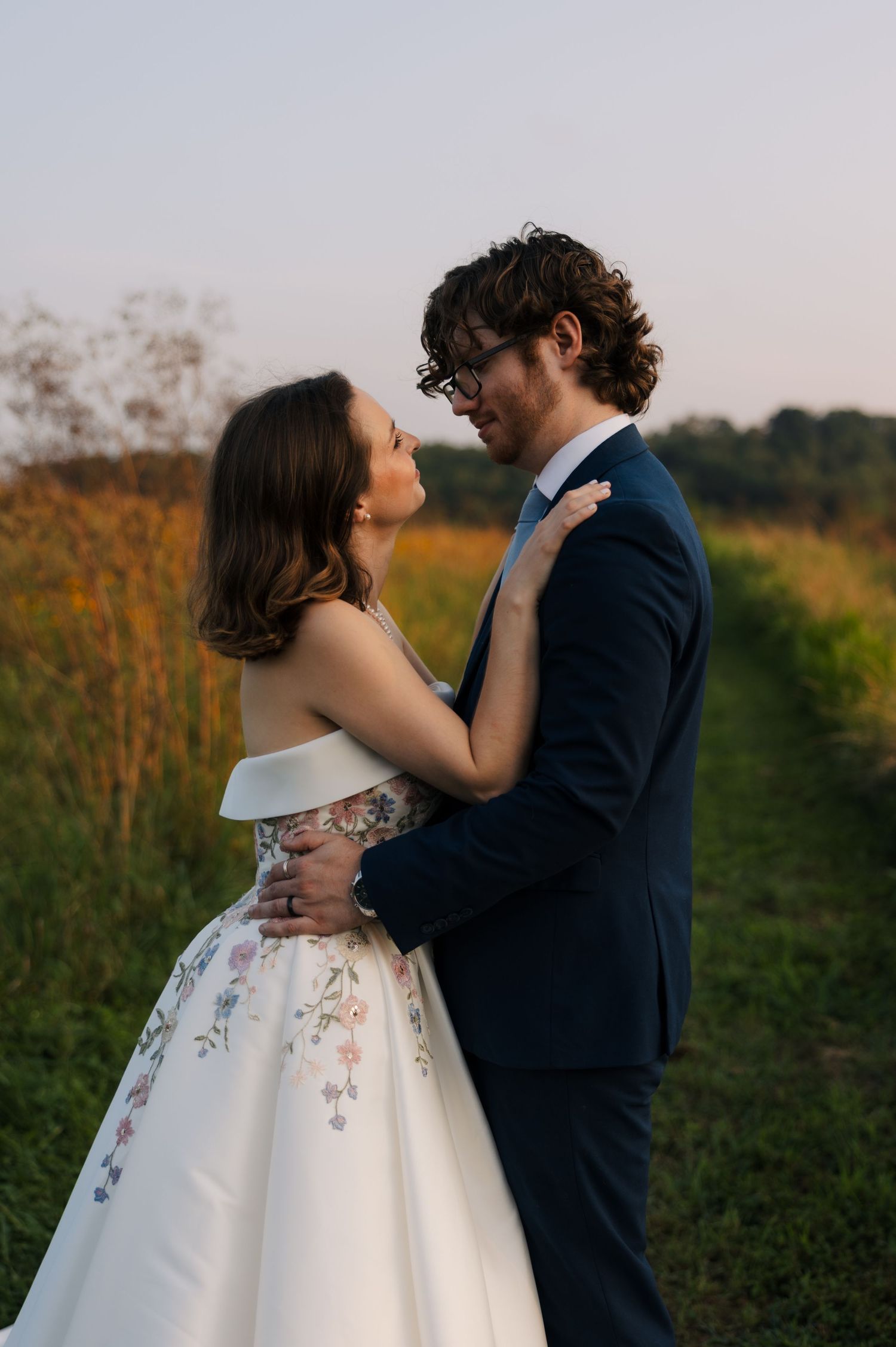 Wedding couple embracing on a field path at sunset, bride in off-shoulder gown.