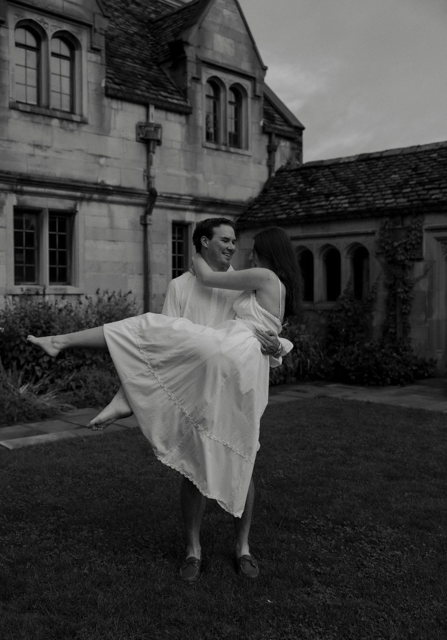 Couple in white sharing a romantic moment, man lifting woman in front of a stone manor house in black and white.
