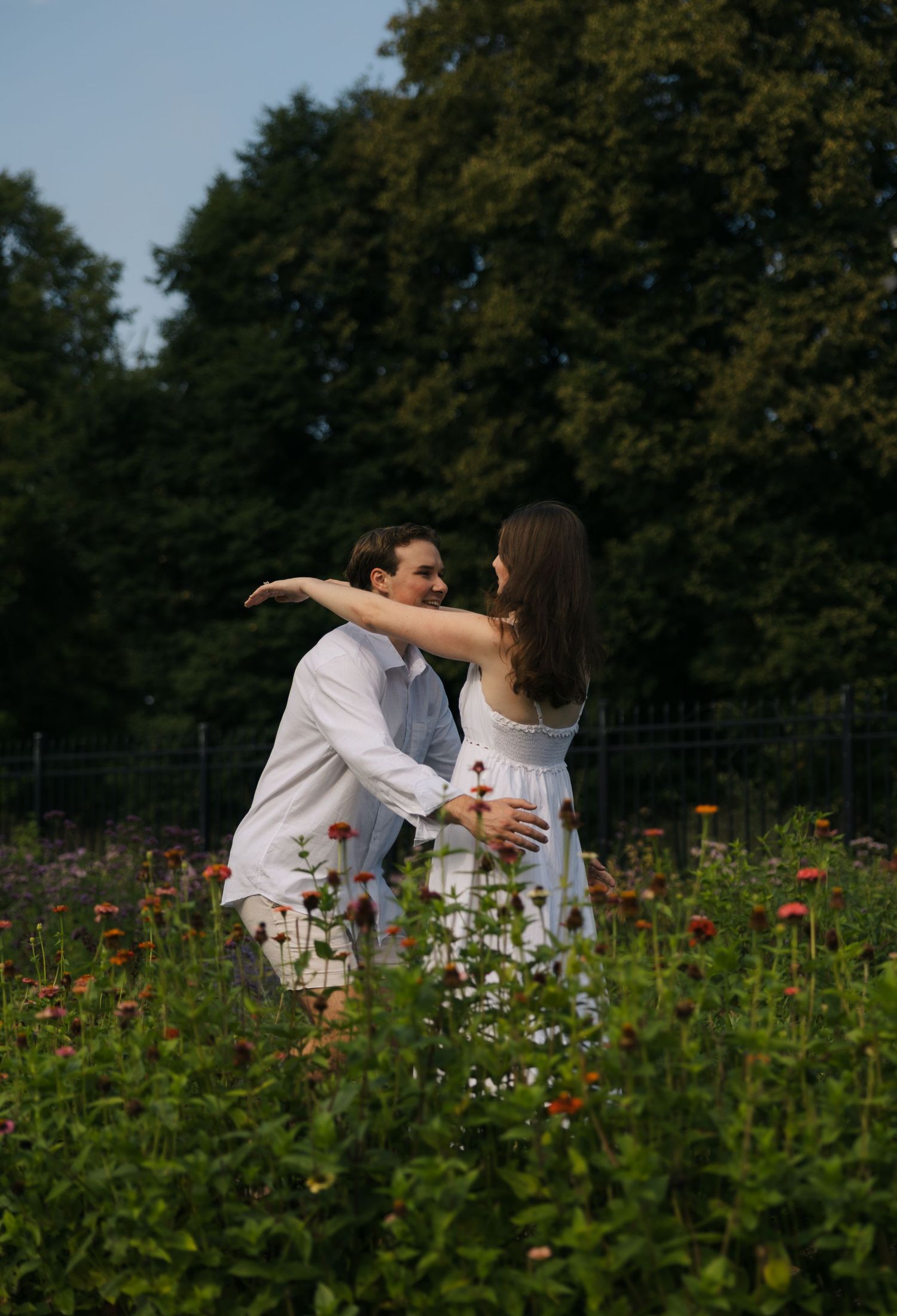 Couple embracing romantically in colorful wildflower garden with tall green trees in background on sunny day.