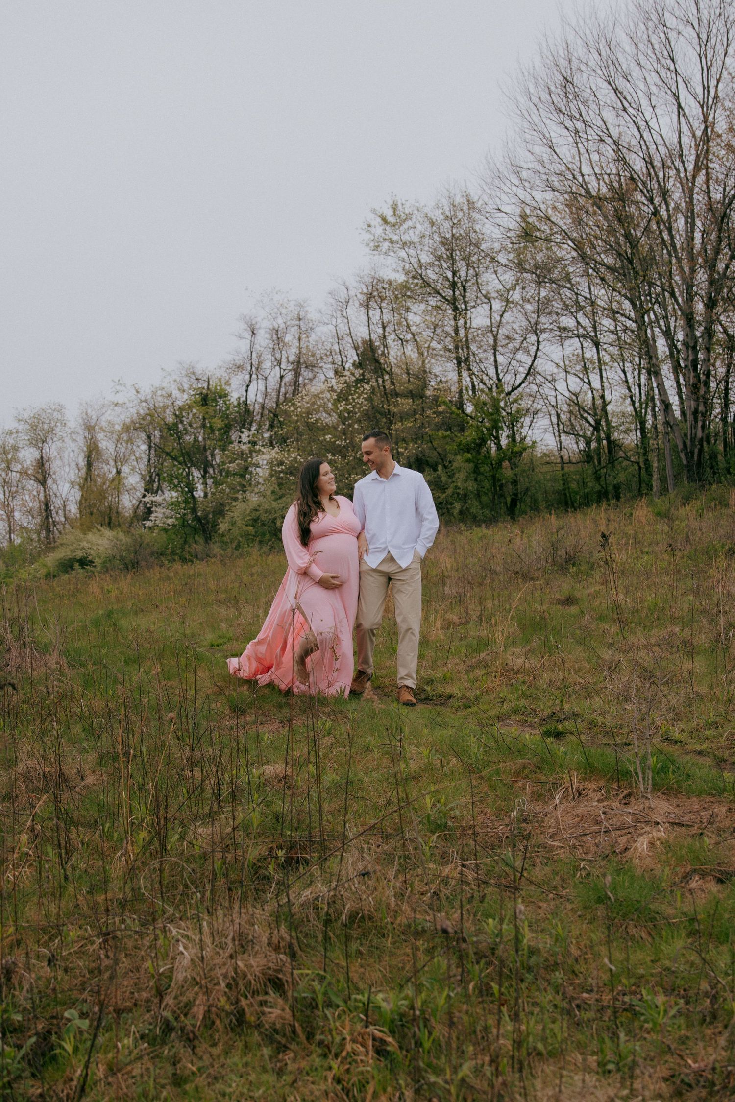 Pregnant couple walking in a green meadow, woman in flowing pink gown, man in white shirt, surrounded by trees.