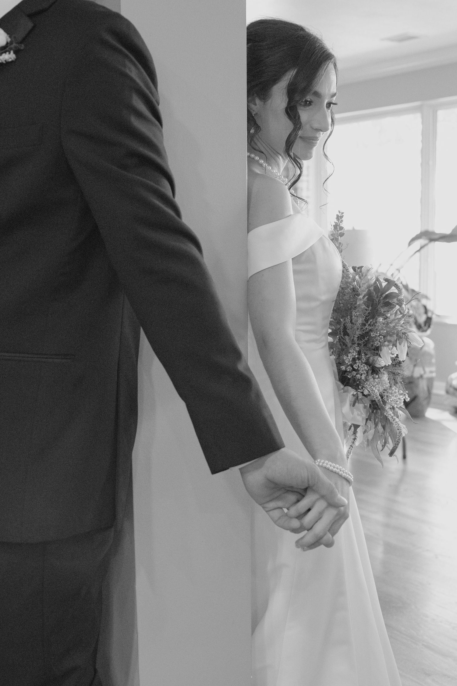 Black and white photo of bride and groom holding hands around a wall, bride holding floral bouquet.