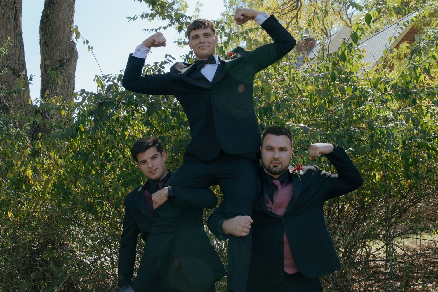Three groomsmen in dark suits strike fun pose with one lifted on shoulders outdoors.