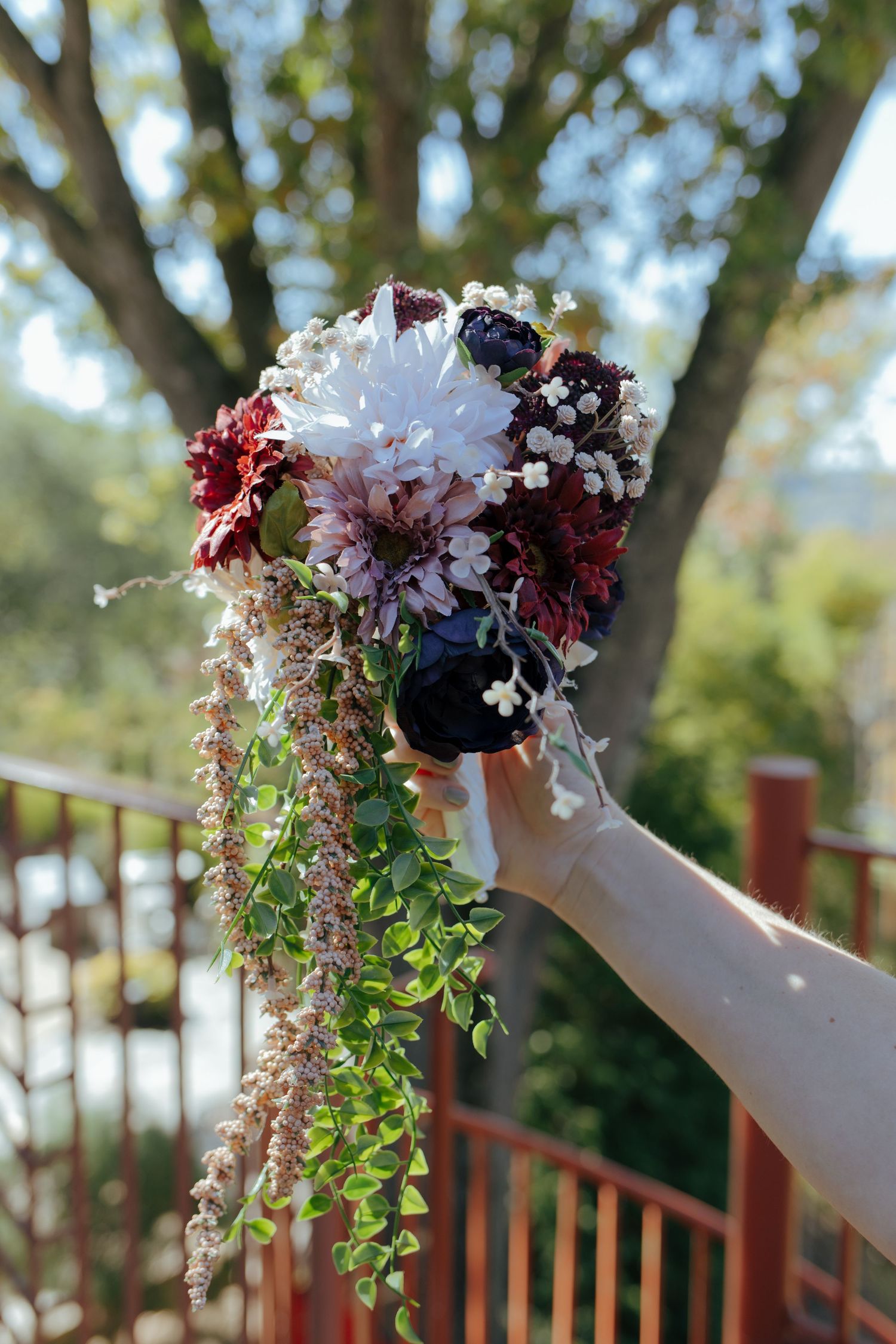 Hand holding a cascading bridal bouquet with burgundy, white, and navy flowers with trailing greenery outdoors.