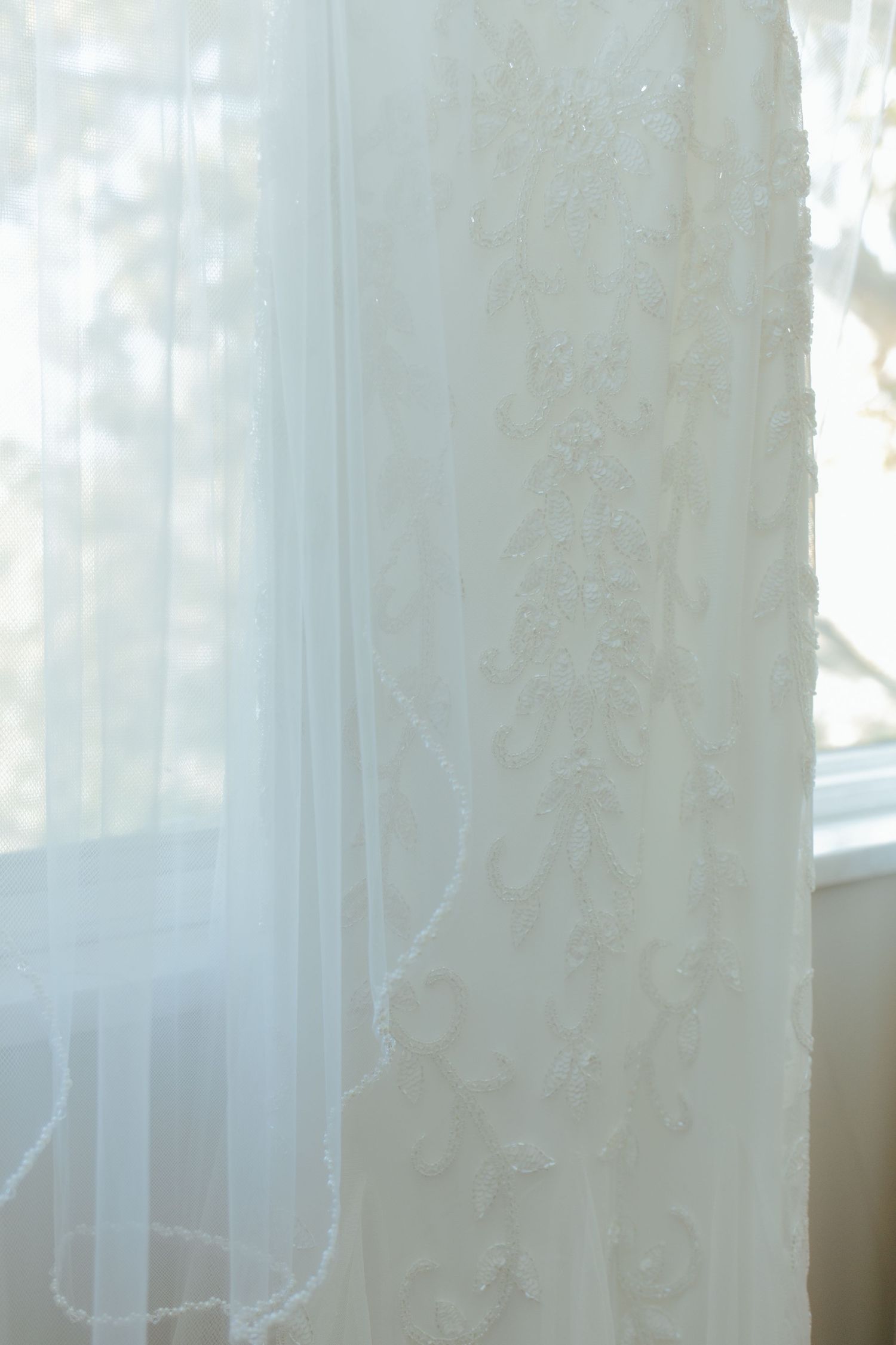 Elegant white lace wedding dress with intricate floral embroidery hanging near a bright window with sheer veil.