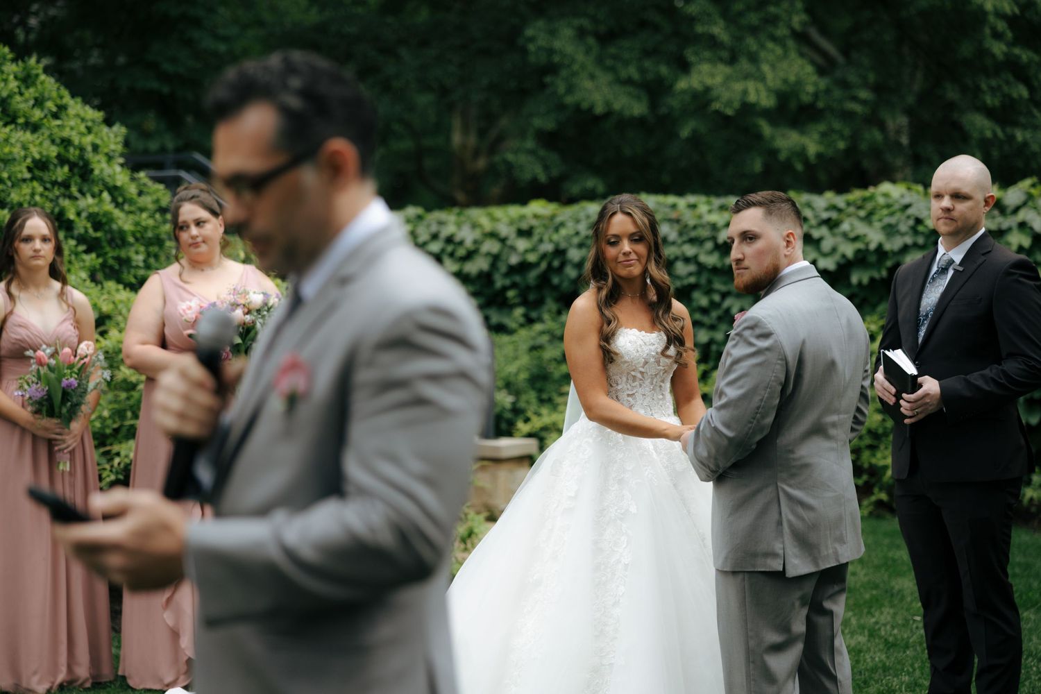 Bride and groom exchange vows outdoors with bridesmaids in pink dresses and lush greenery at elegant garden wedding.