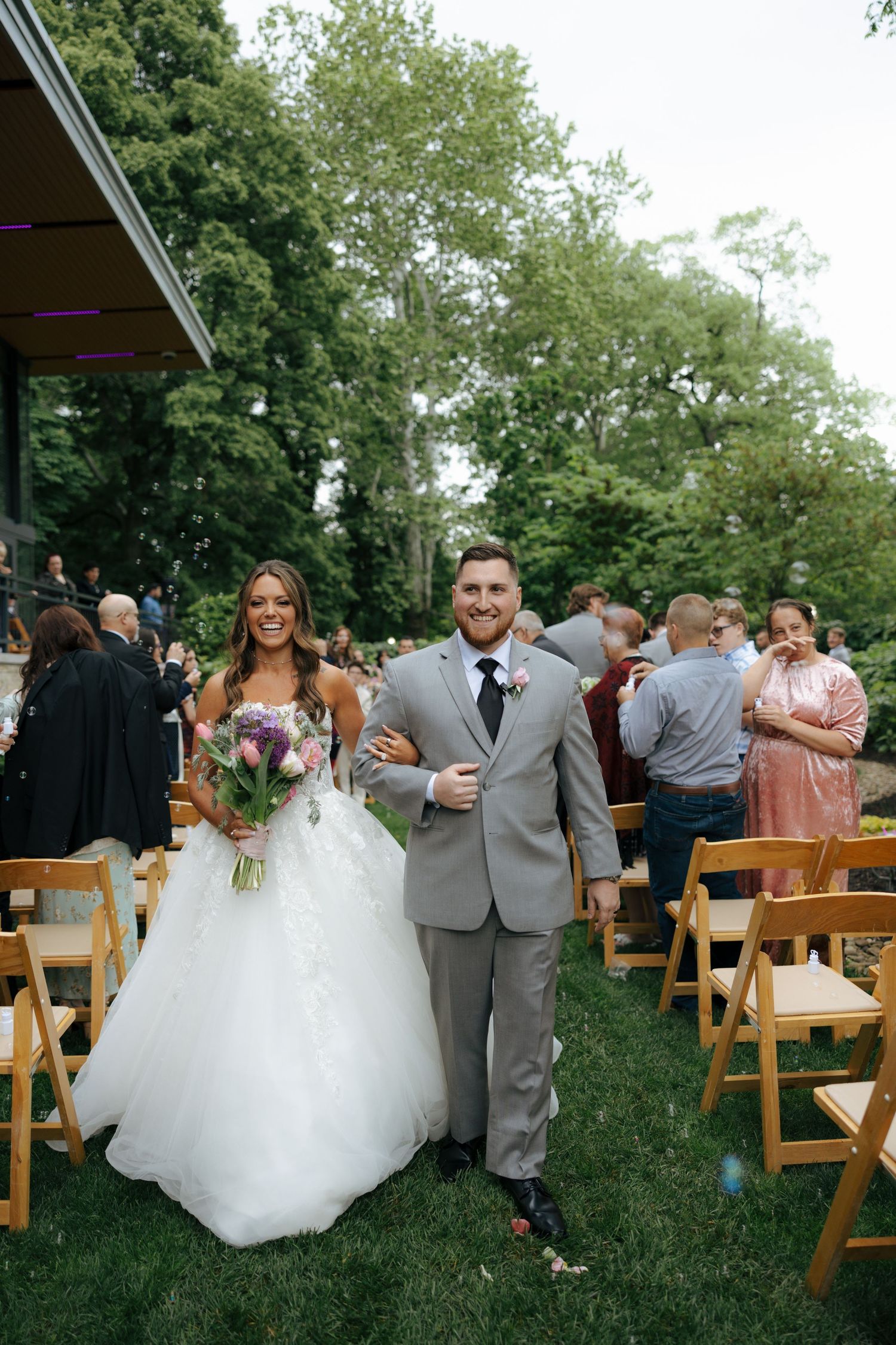 Joyful newlywed couple exits outdoor wedding ceremony aisle, bride in lace gown with floral bouquet.
