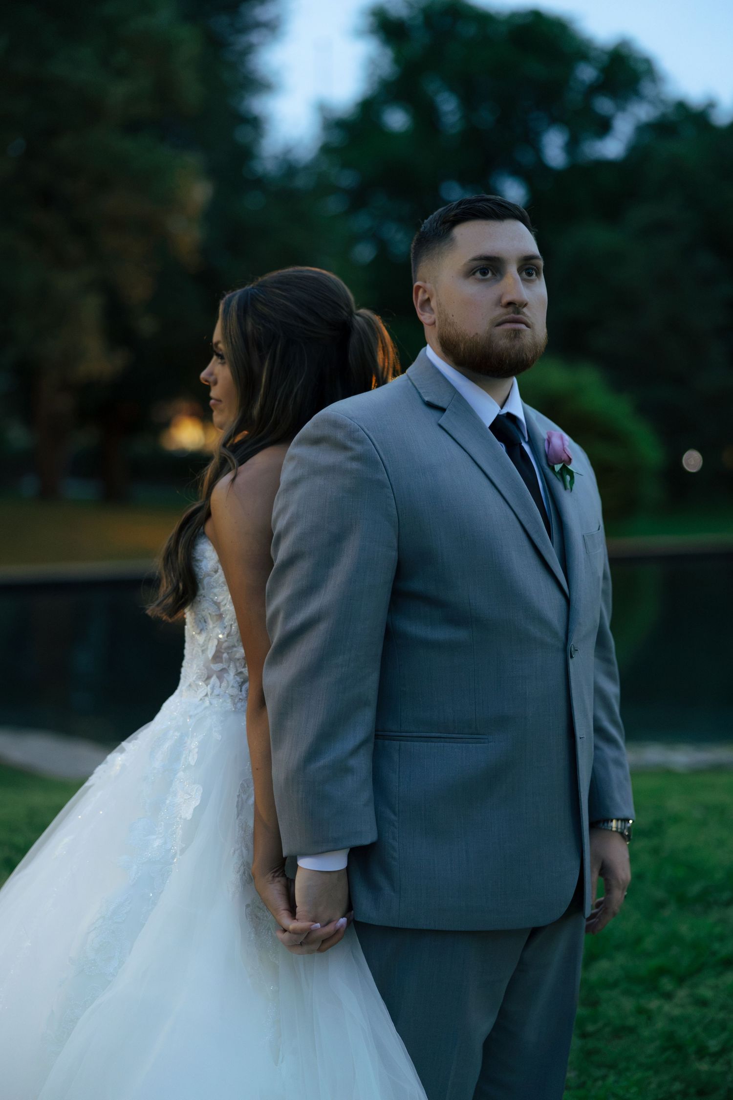 Groom in gray suit stands back to back with bride in lace ballgown near a serene pond at dusk.