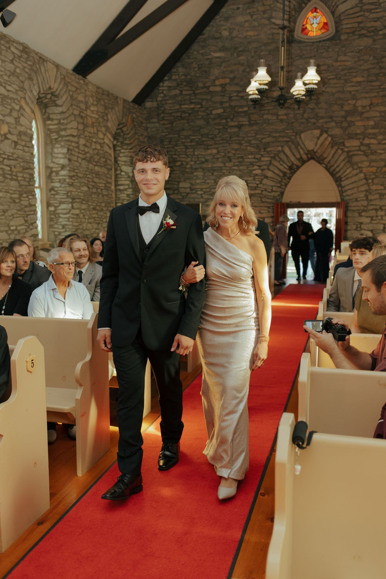 Young man in tuxedo escorts elegantly dressed woman down red carpet aisle in stone church wedding ceremony.