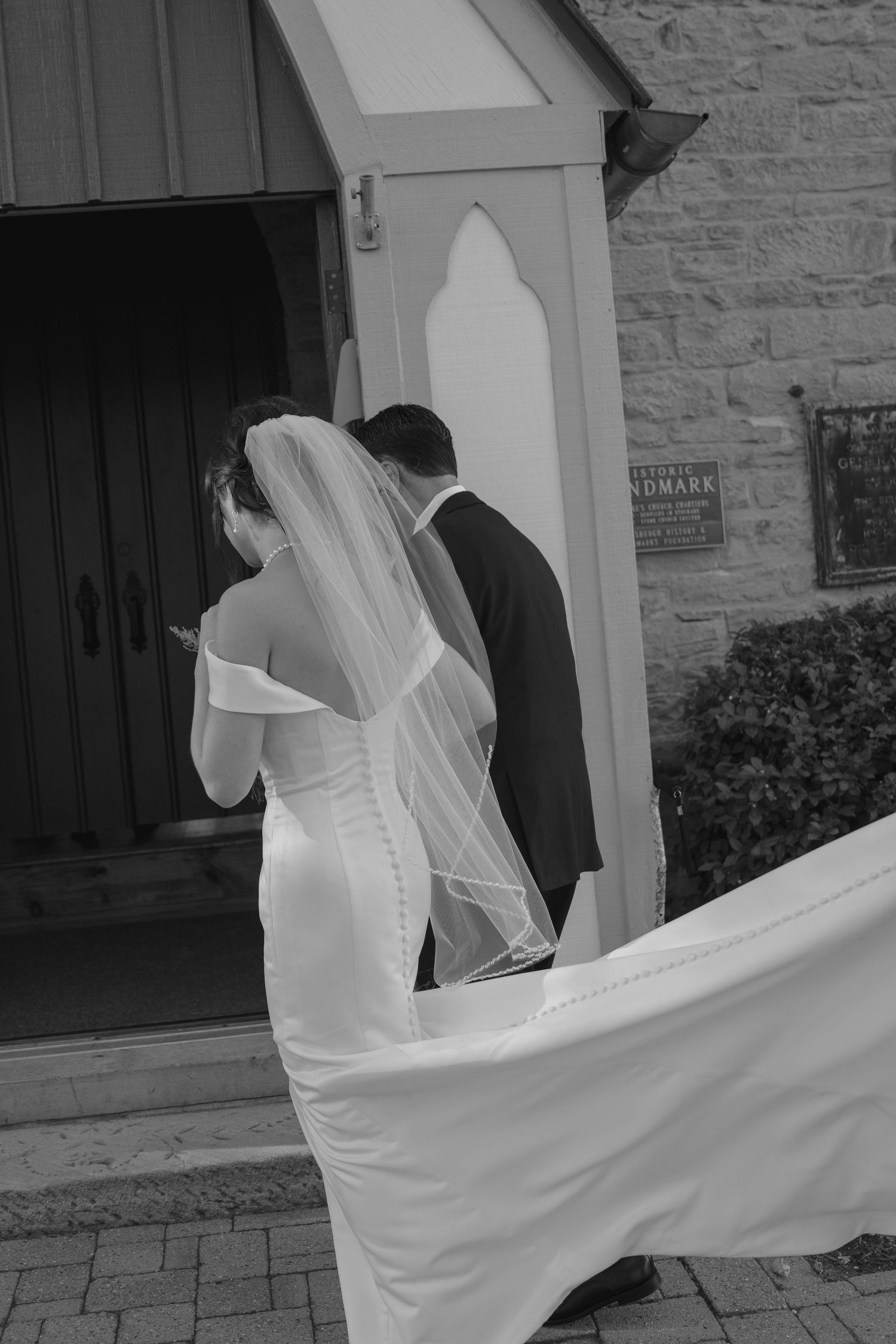 Black and white photo of bride and groom entering a stone church, wedding veil flowing in the breeze.