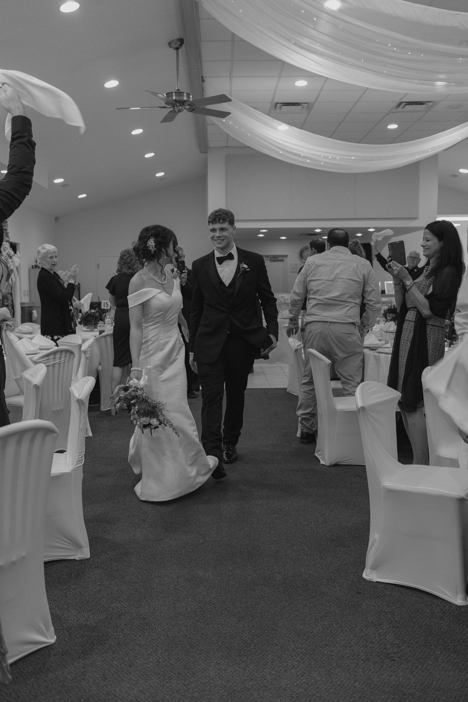 Bride and groom walking down reception aisle while guests celebrate in black and white photo.