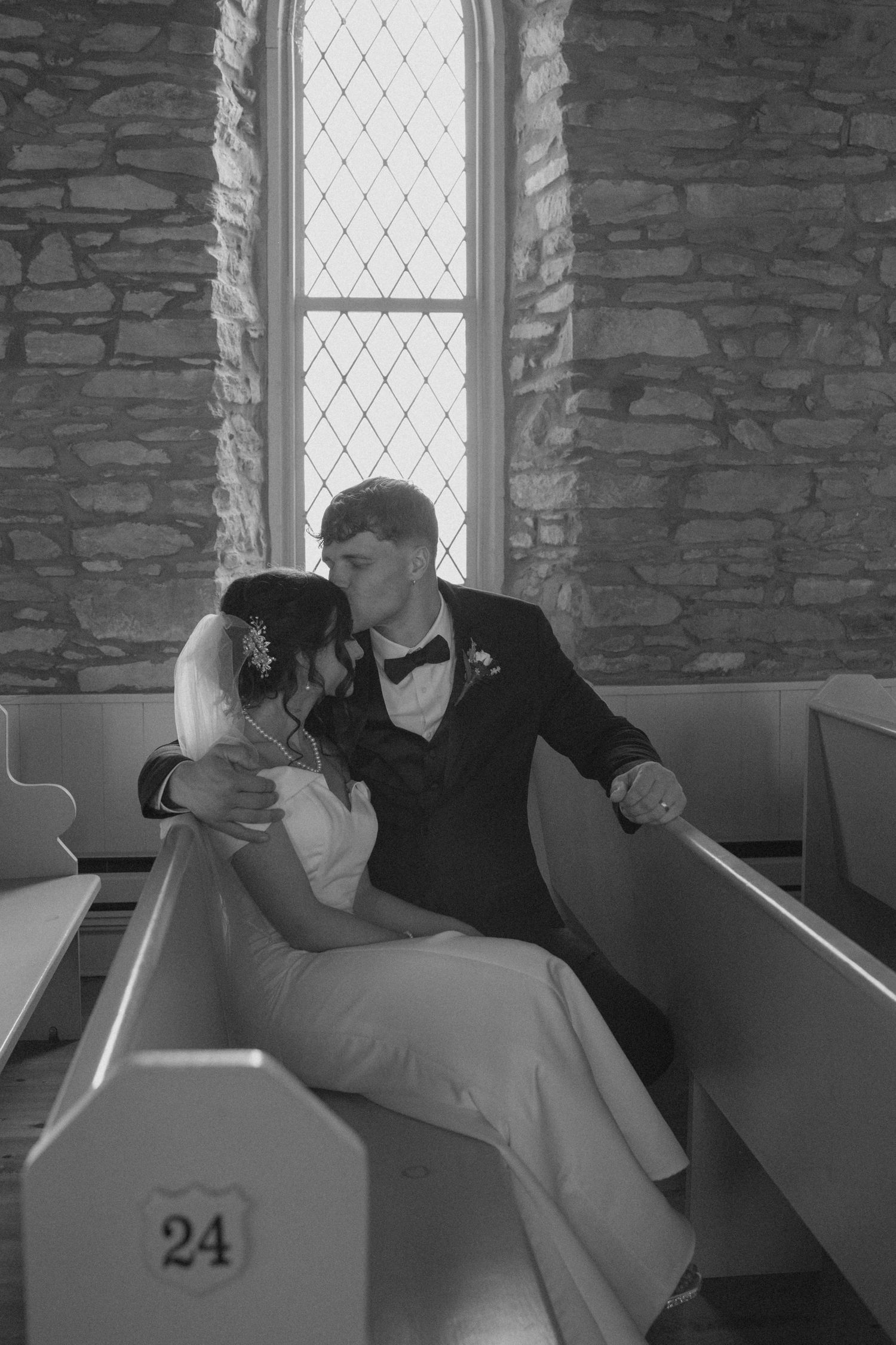 Black and white photo of bride and groom kissing on pew 24 inside a historic stone church.