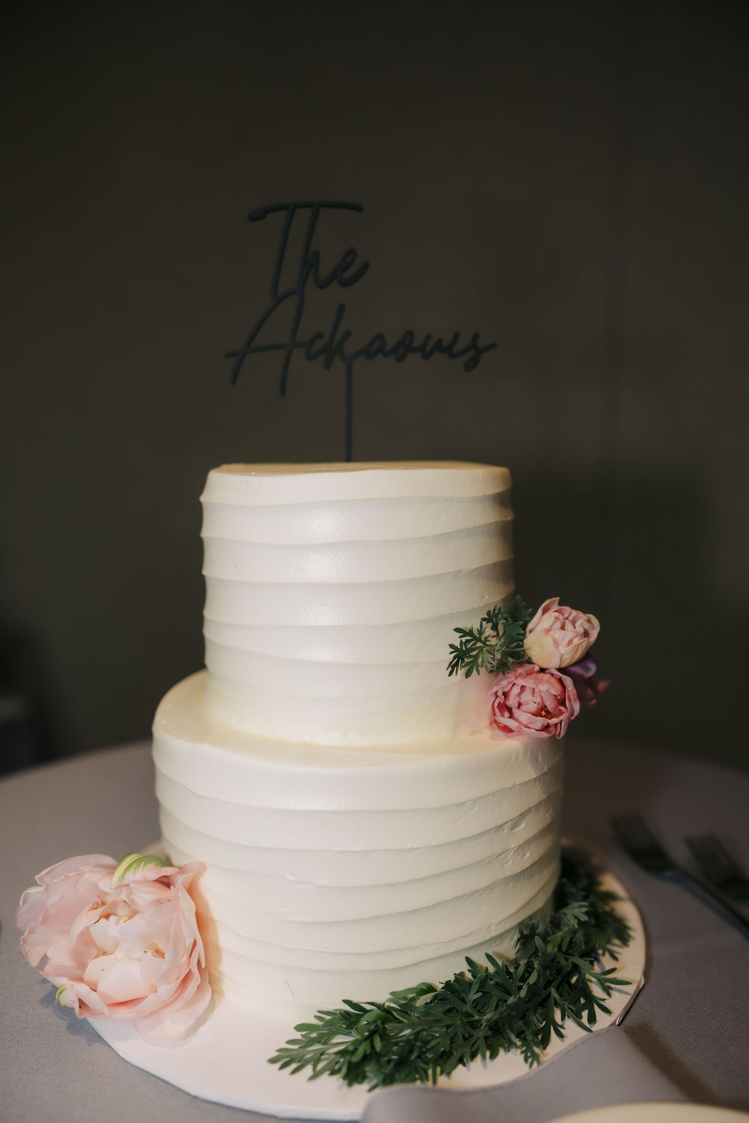 Two-tier white textured wedding cake with The Adkaoves topper, adorned with pink roses and greenery.