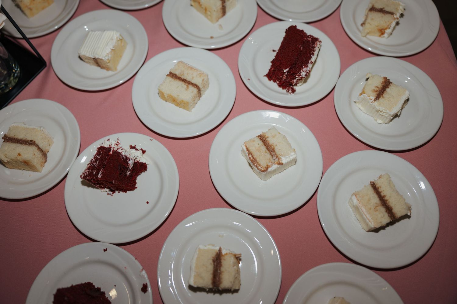 Assorted slices of white and red velvet cake served on white plates arranged on a pink tablecloth.