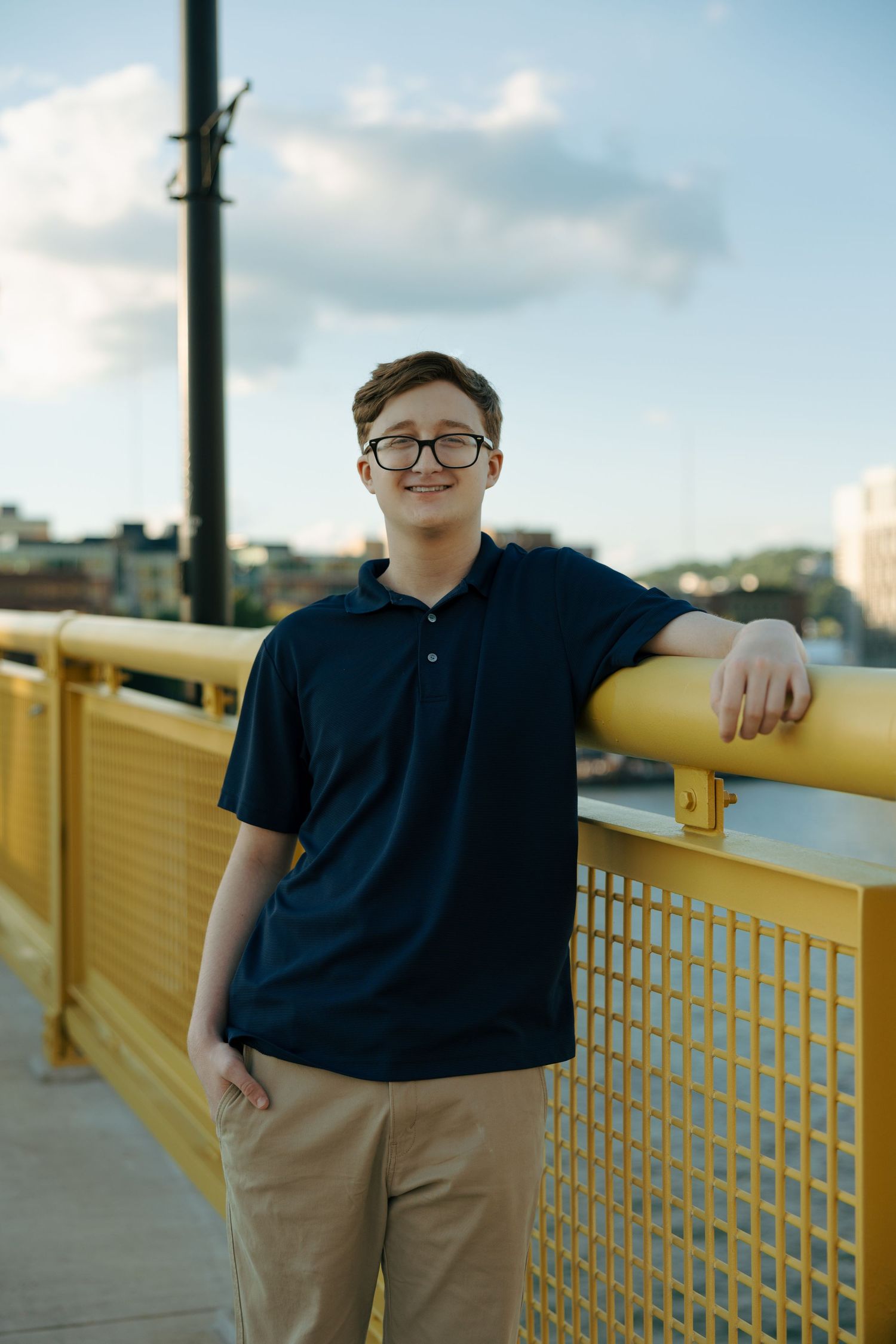 Young man in navy polo and glasses leaning on yellow bridge railing with city skyline behind him.