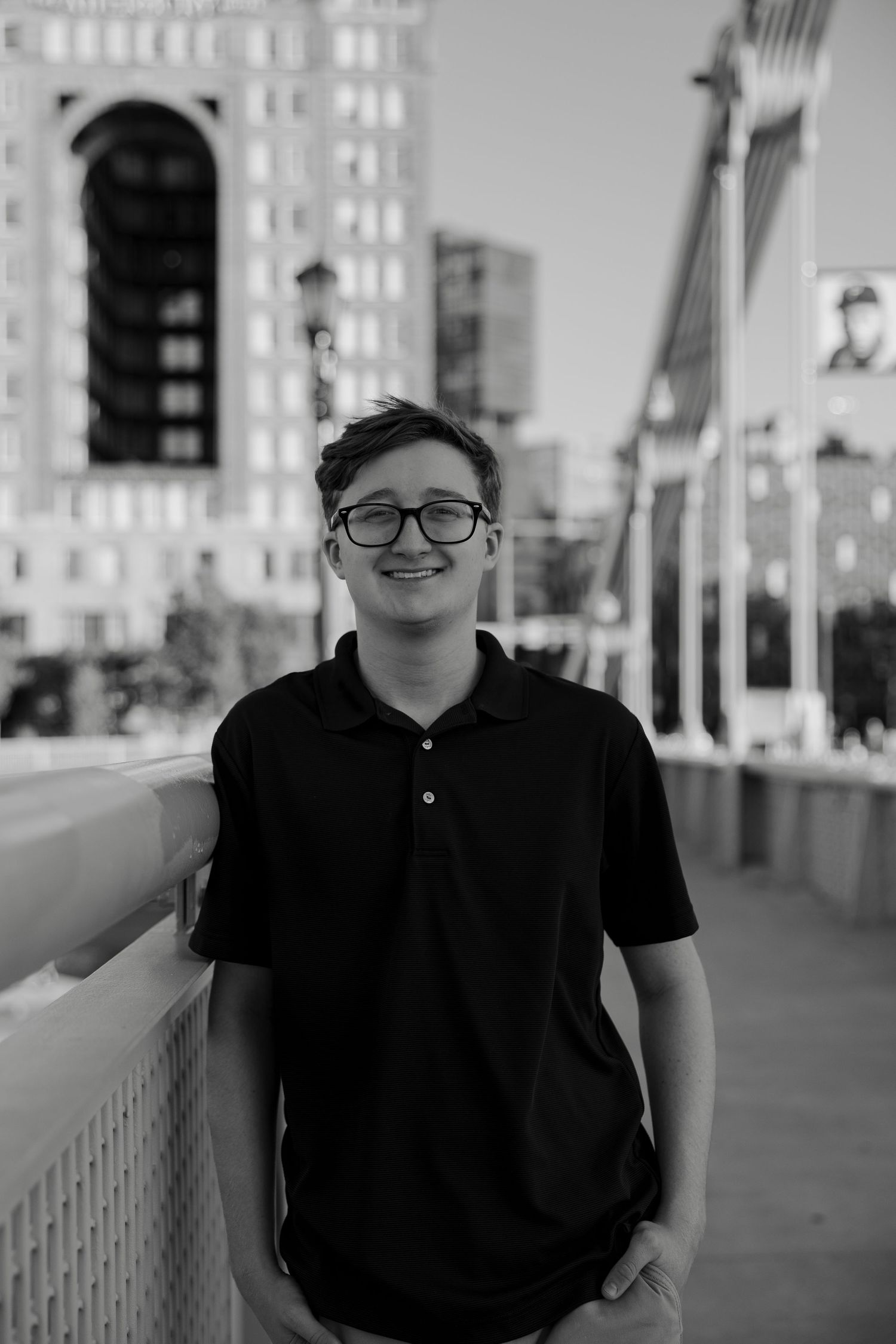 Smiling teen boy with glasses in black polo shirt leaning on bridge railing with urban skyline in black and white.