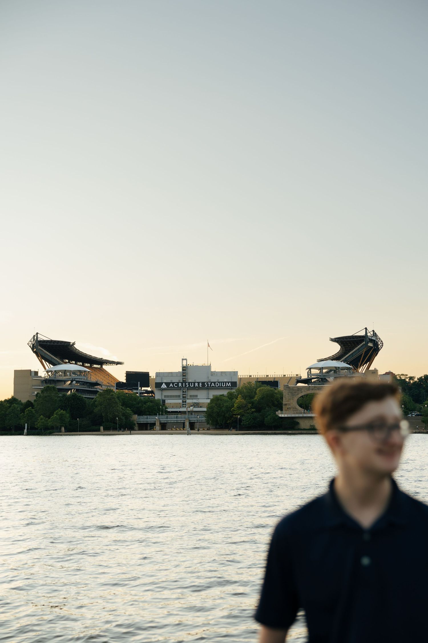 Teen boy in navy polo standing near a river with Acrisure Stadium visible across the water at dusk.