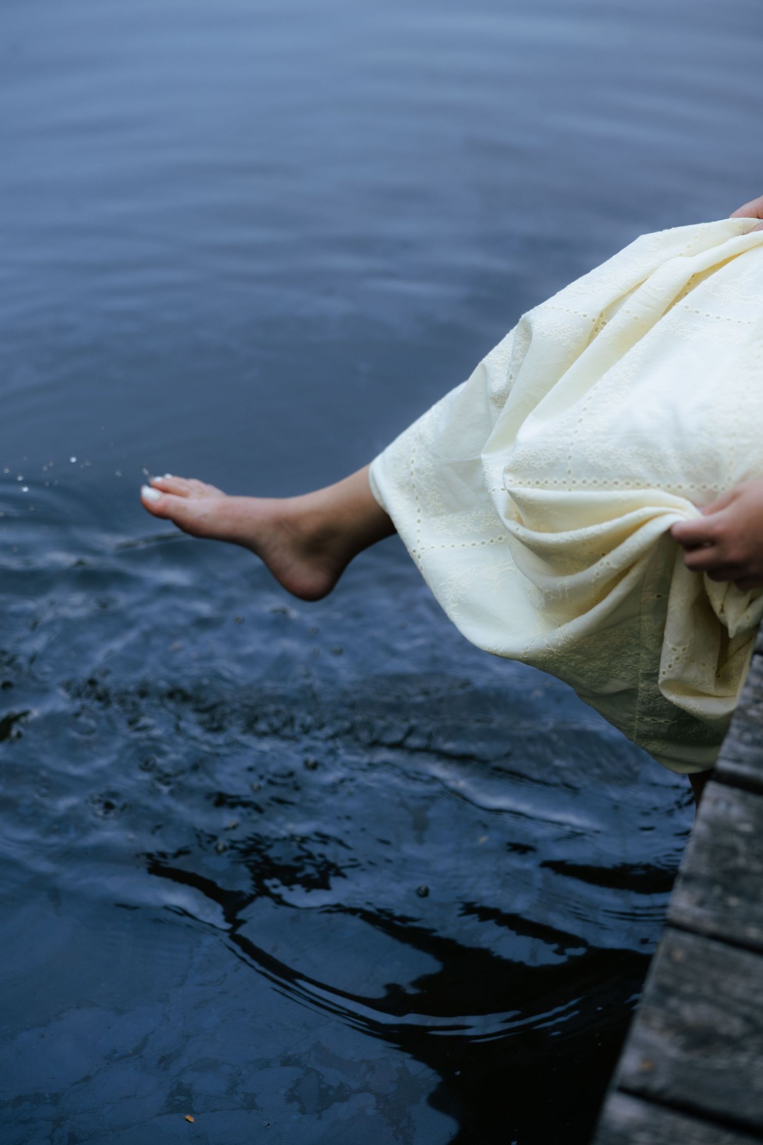 Bare feet of a woman in a cream dress dangling over still water from a rustic wooden dock.