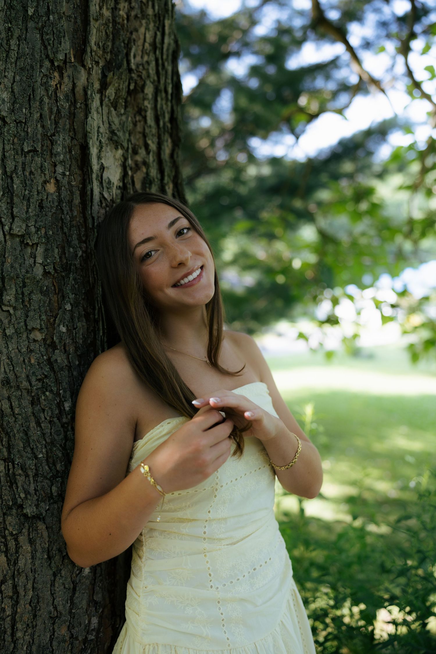 Young woman in white strapless dress smiling outdoors leaning against tree trunk in lush green park.