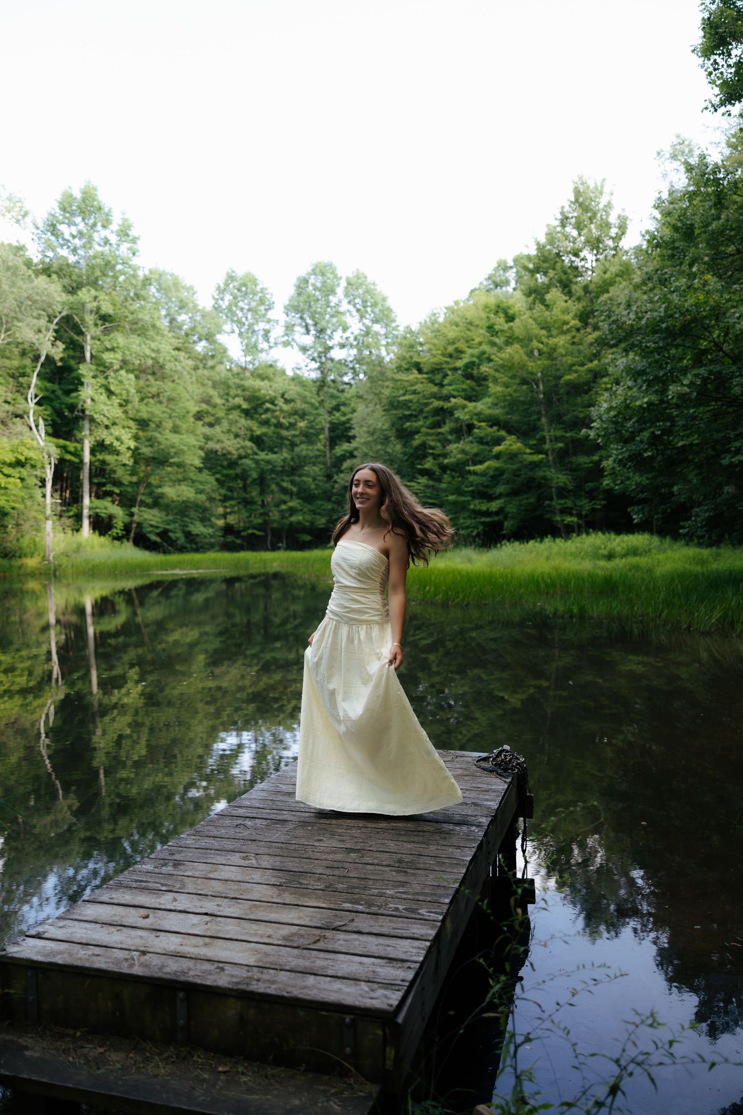 Young woman in cream strapless gown standing on wooden dock by reflective forest lake.