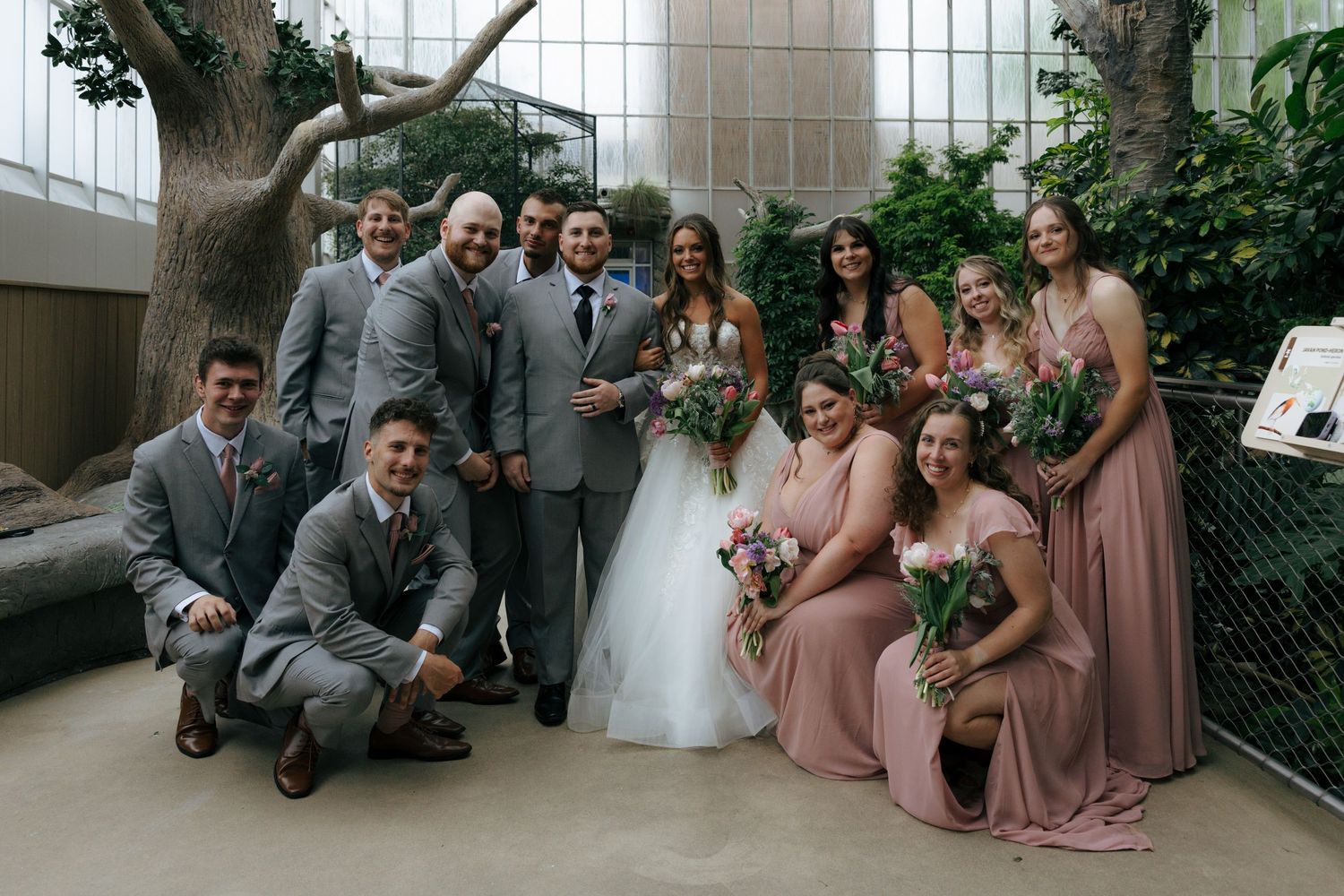 Wedding party poses indoors, groomsmen in gray suits and bridesmaids in dusty rose gowns surround smiling bride and groom.