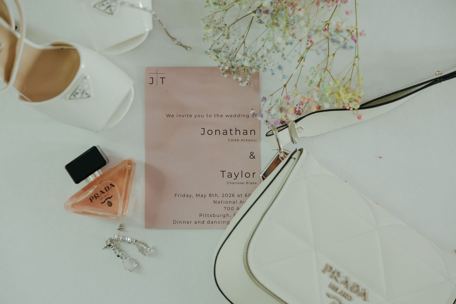 Wedding flat lay featuring a pink invitation for Jonathan and Taylor, Prada perfume, white heels, and floral accents.