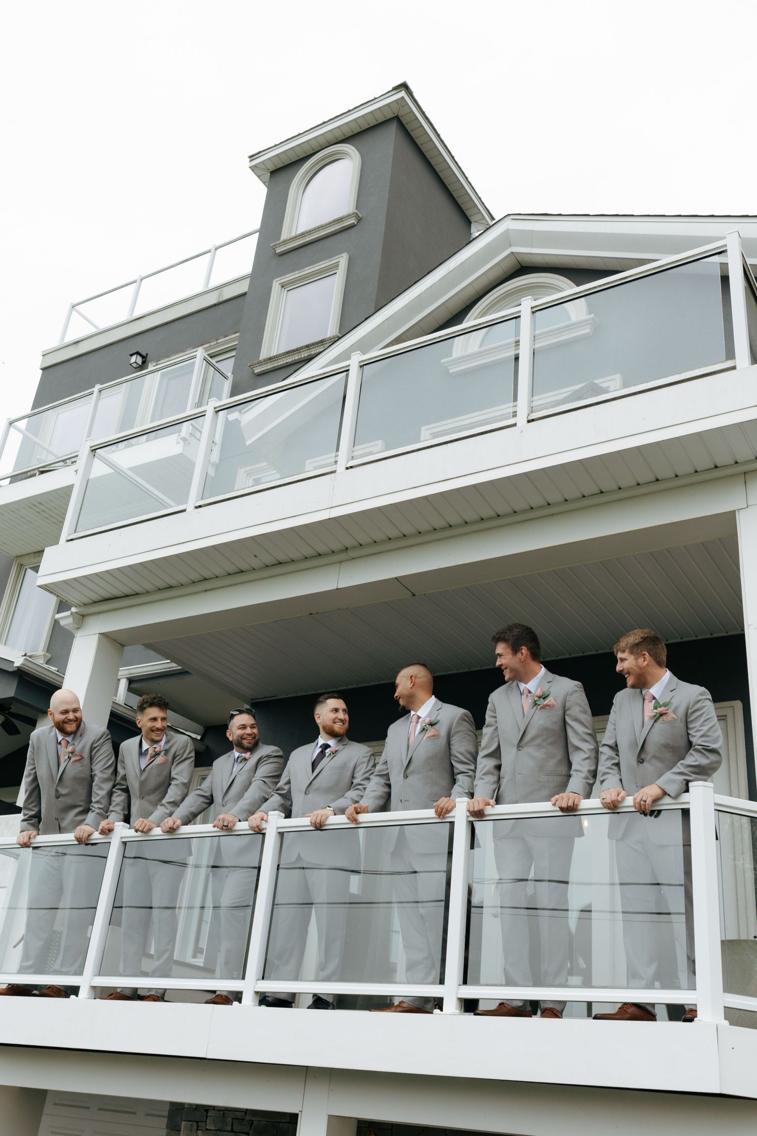 Seven groomsmen in gray suits stand together on a white balcony railing of a modern coastal wedding venue.