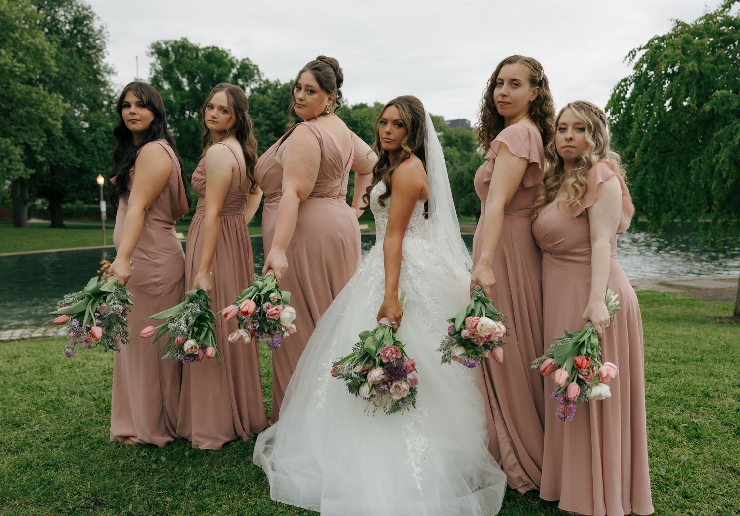Bride in white gown poses with five bridesmaids in dusty rose dresses holding floral bouquets outdoors.