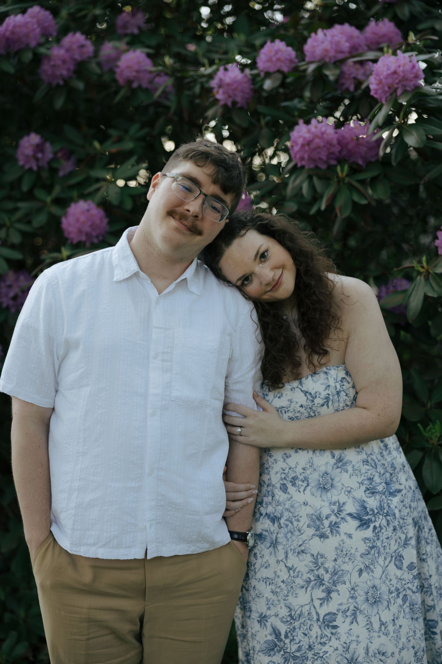 Engaged couple leaning together affectionately in front of blooming purple rhododendron bushes.