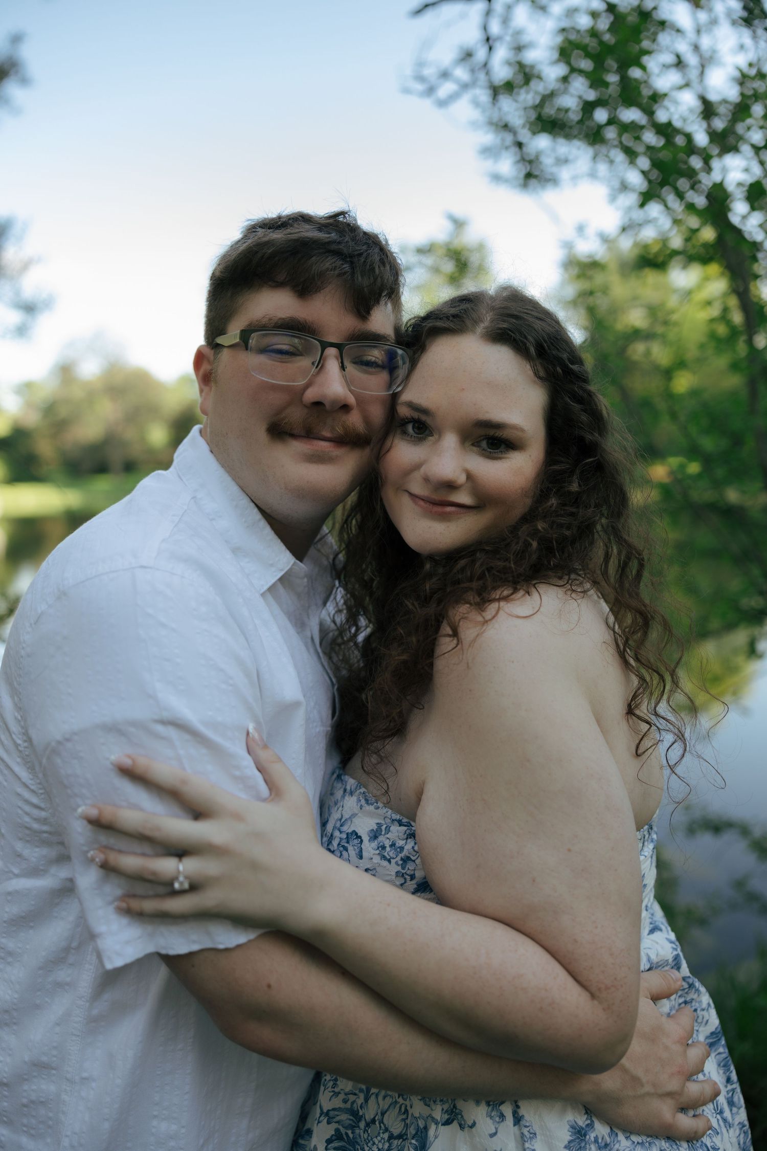Happy engaged couple embracing and smiling near a pond surrounded by lush green trees.