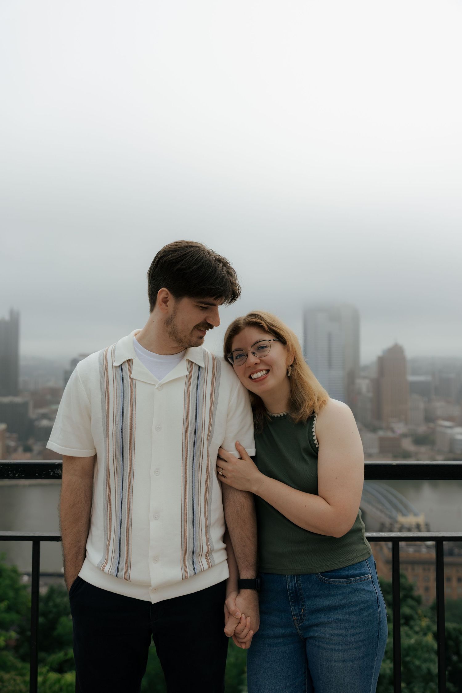 A couple holds hands and smiles on a rooftop overlooking a city skyline on a cloudy day.