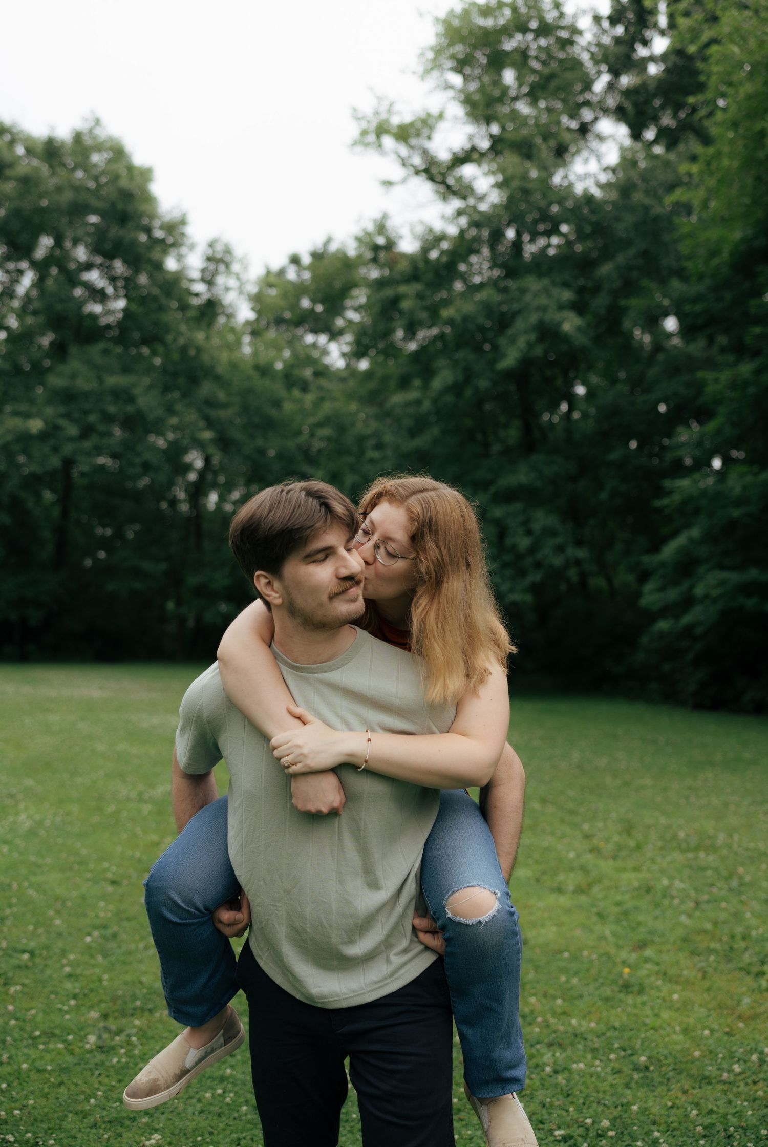 Woman kisses man on the cheek while he gives her a piggyback ride in a green outdoor park.