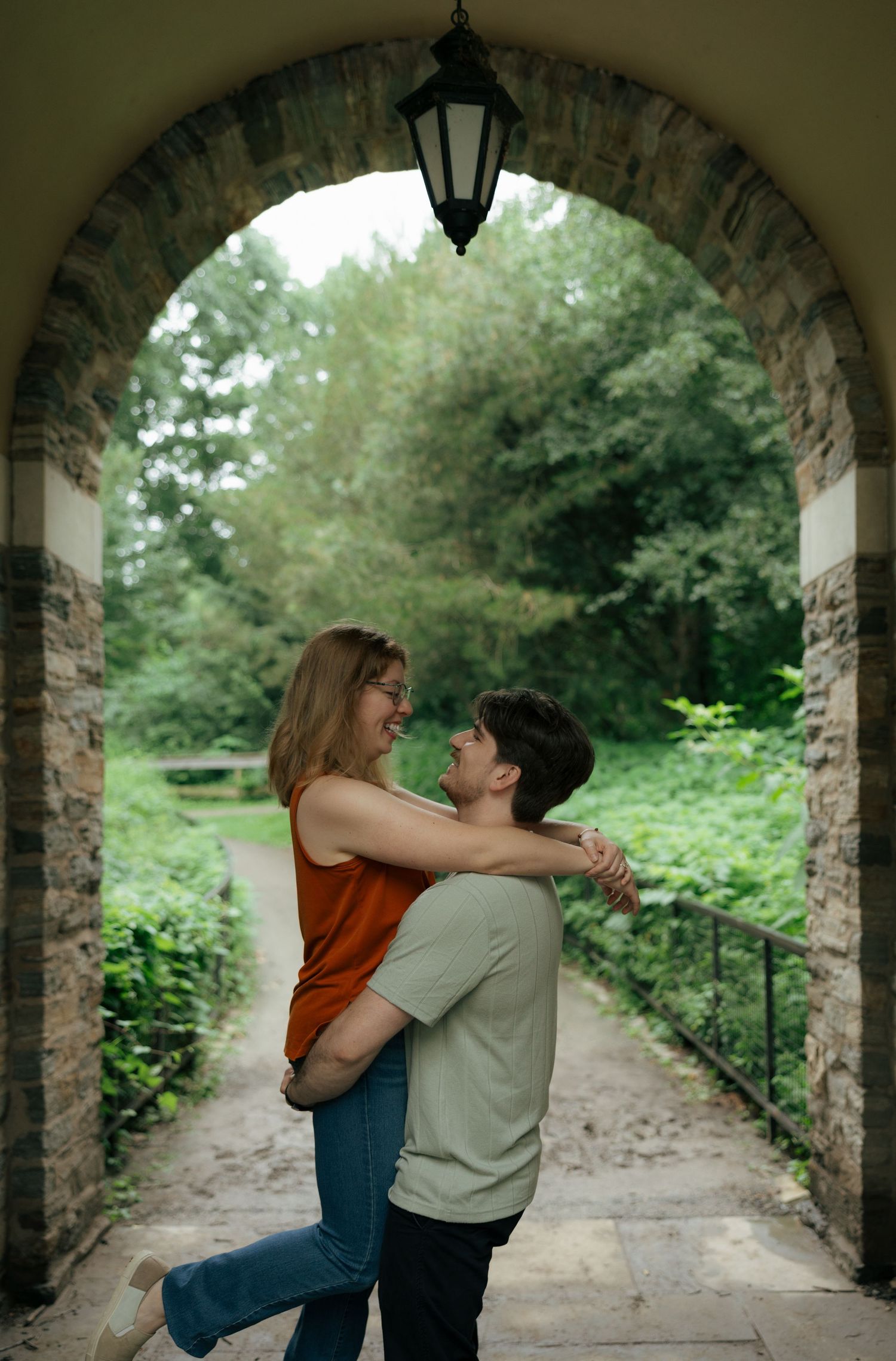A joyful couple embraces and spins under a stone arch with a garden path behind them.