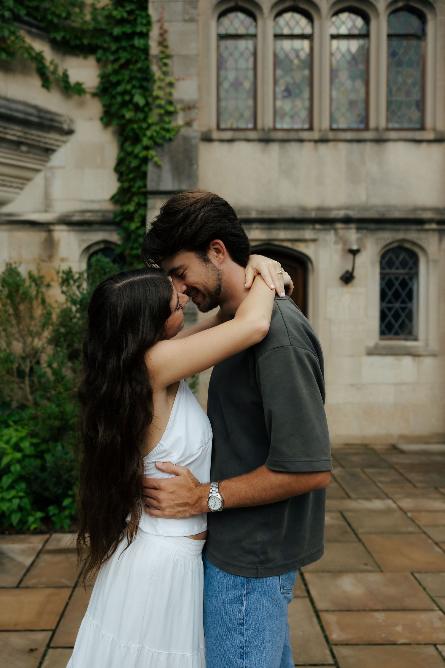 Couple embracing and smiling at each other outside a stone building with ivy and gothic windows.