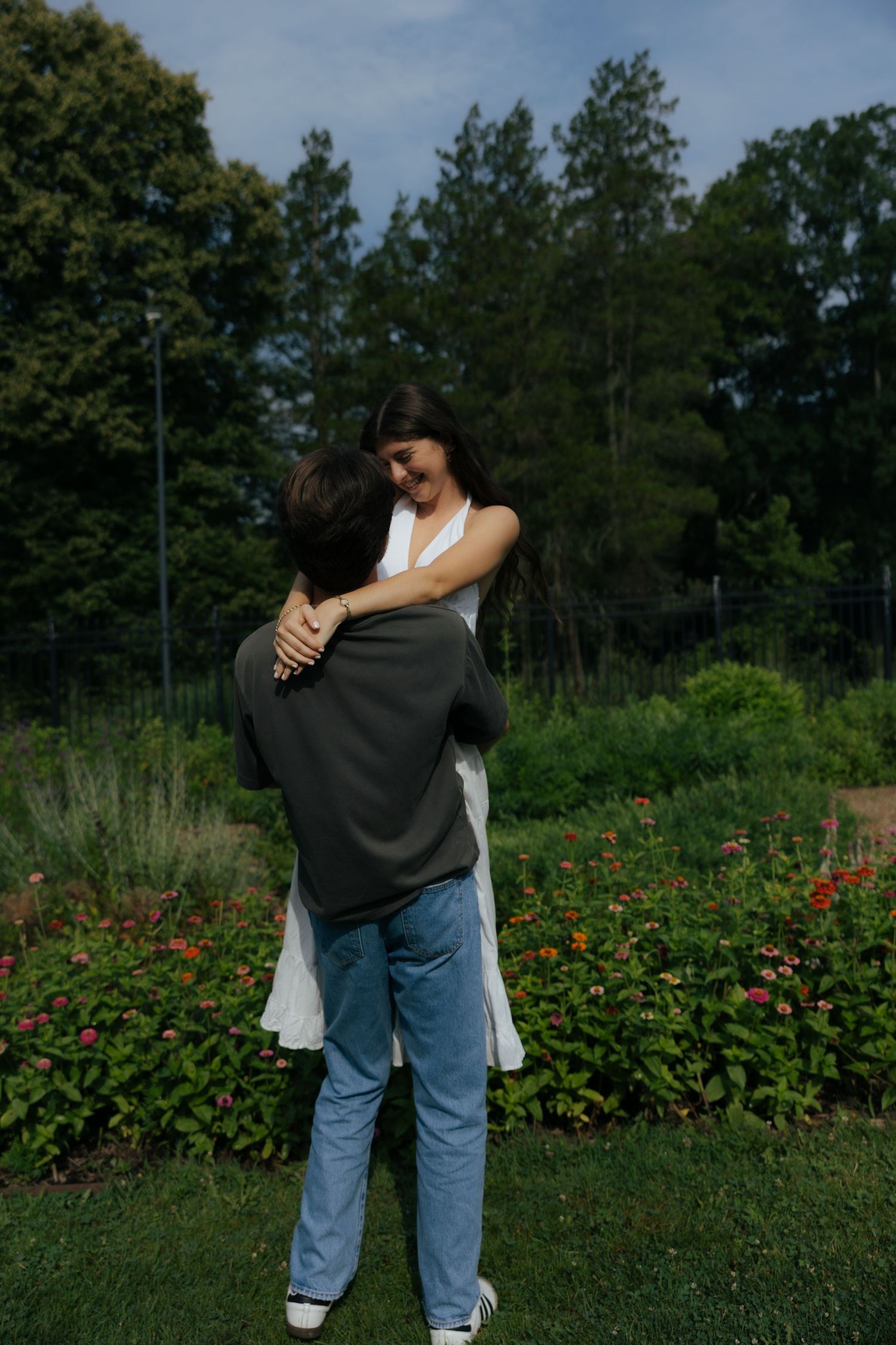 Man lifting woman in white dress as she smiles and wraps her arms around him in a flower garden.
