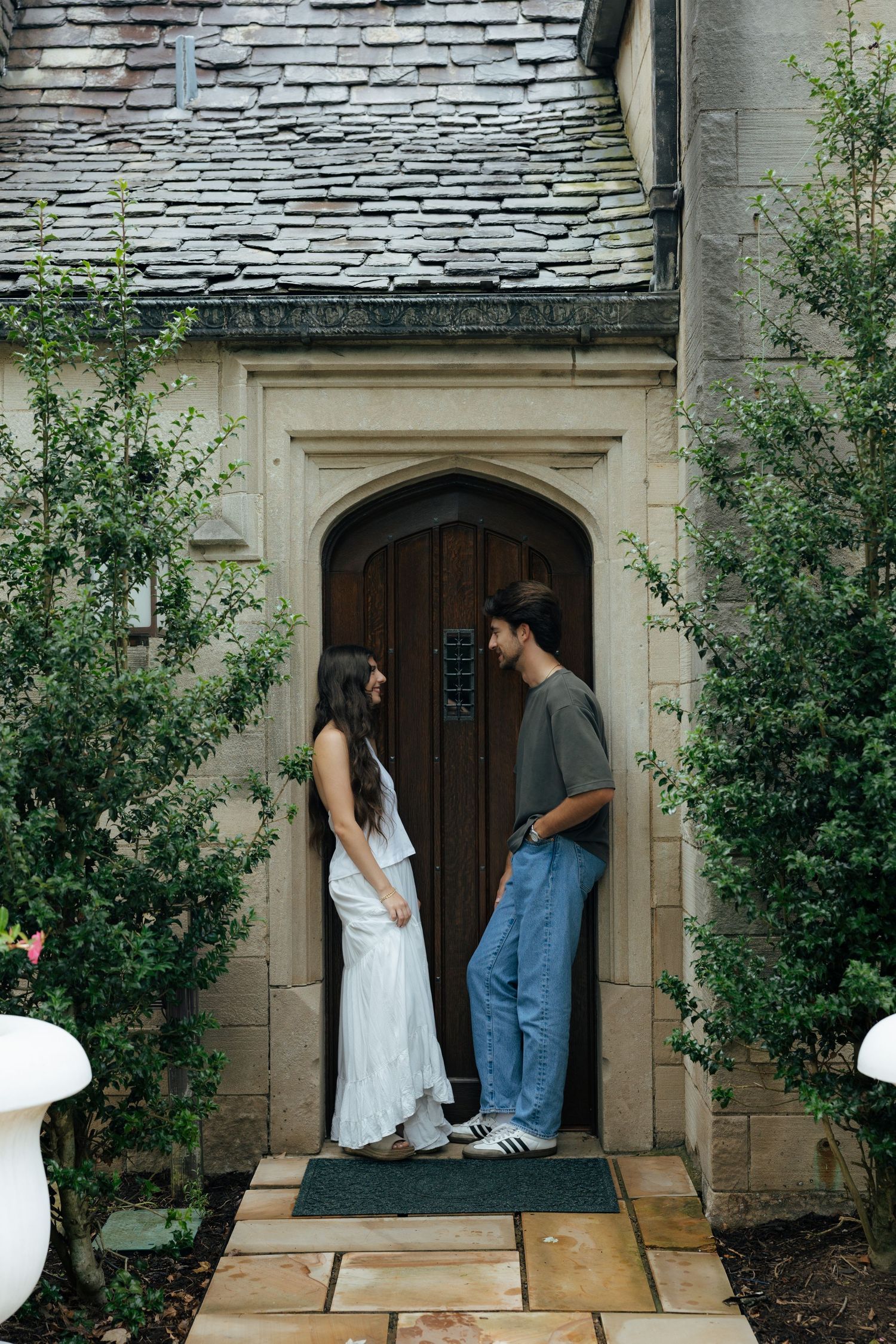 Couple standing face to face at an arched wooden door of a stone building surrounded by lush greenery.