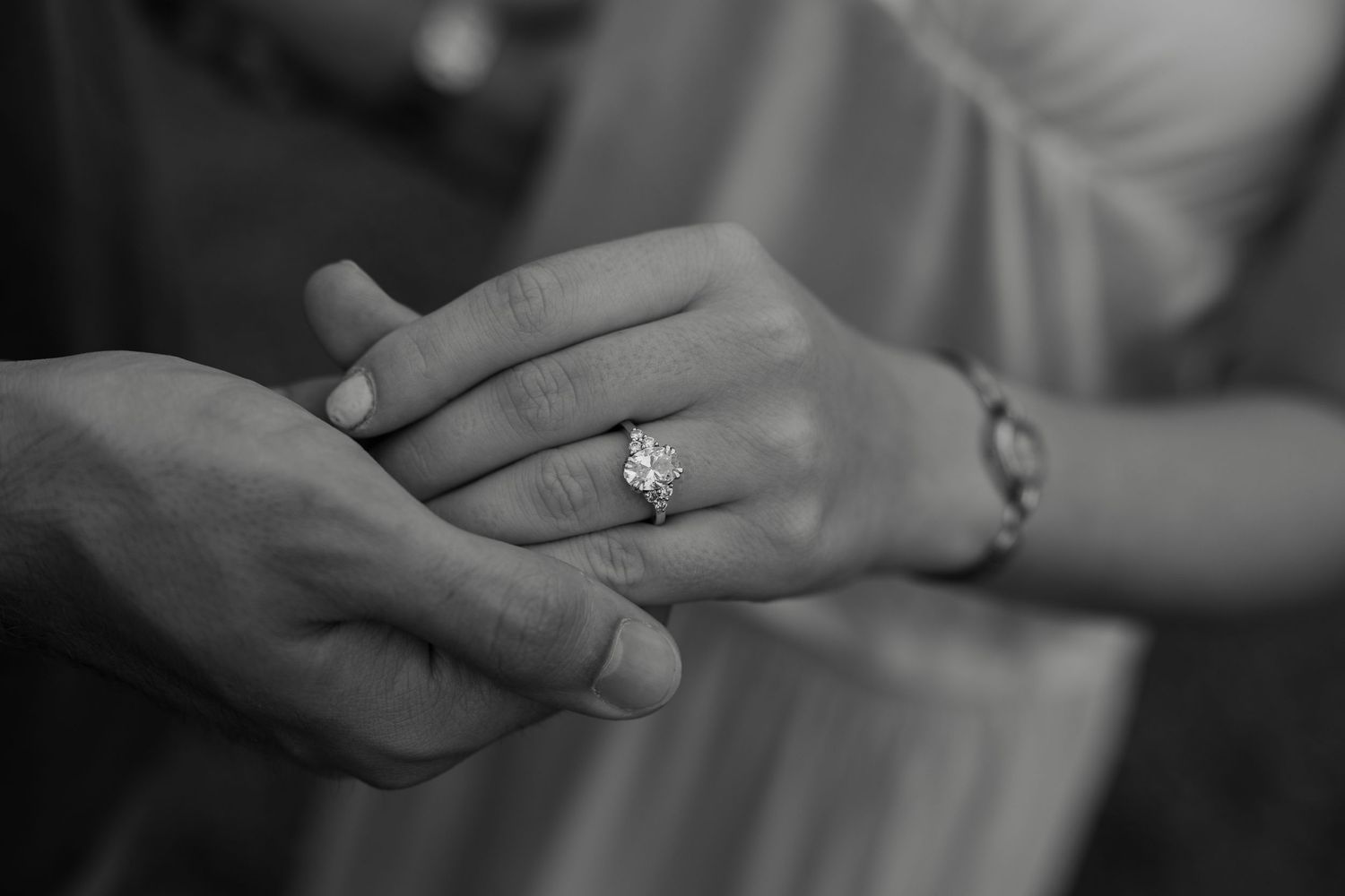 Black and white photo of a couple holding hands, showcasing a diamond engagement ring on the woman's finger.