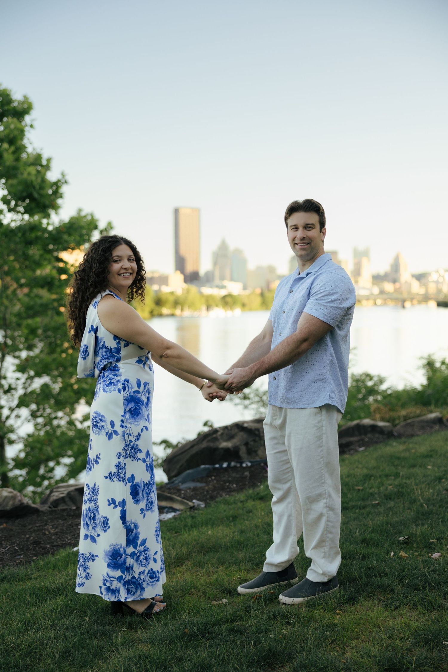 Couple holding hands outdoors by a river with city skyline, woman in floral dress and man in blue shirt.