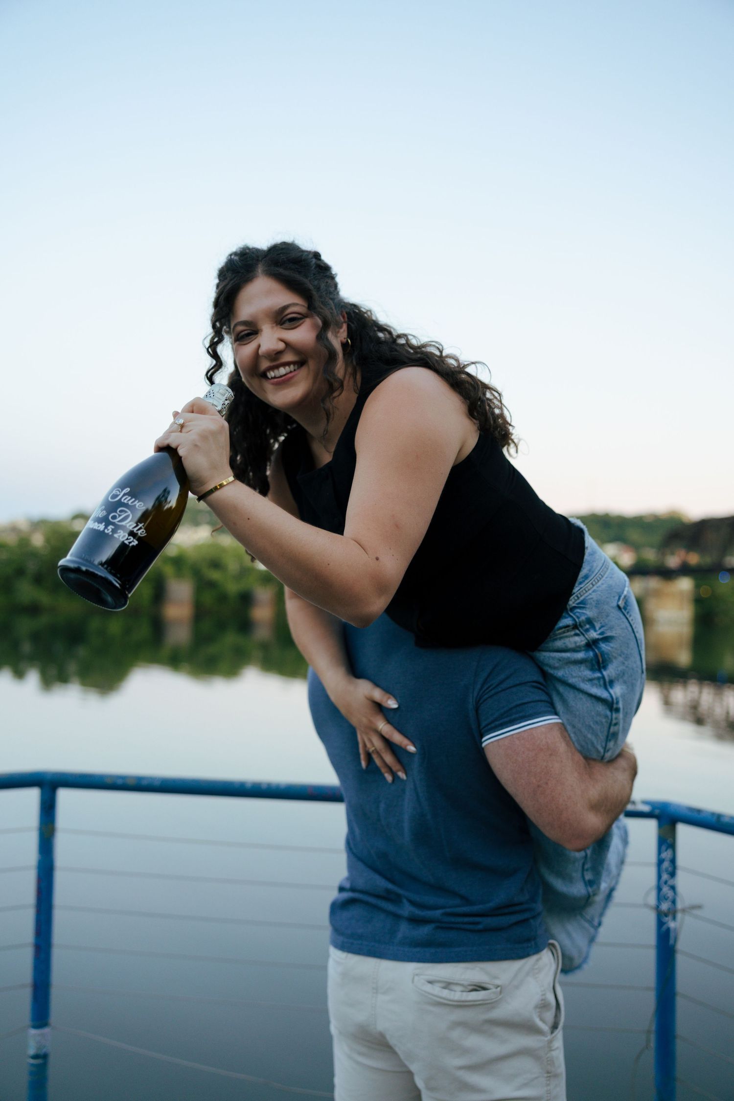 Happy couple celebrating engagement with champagne bottle on waterfront deck at dusk, woman laughing joyfully.