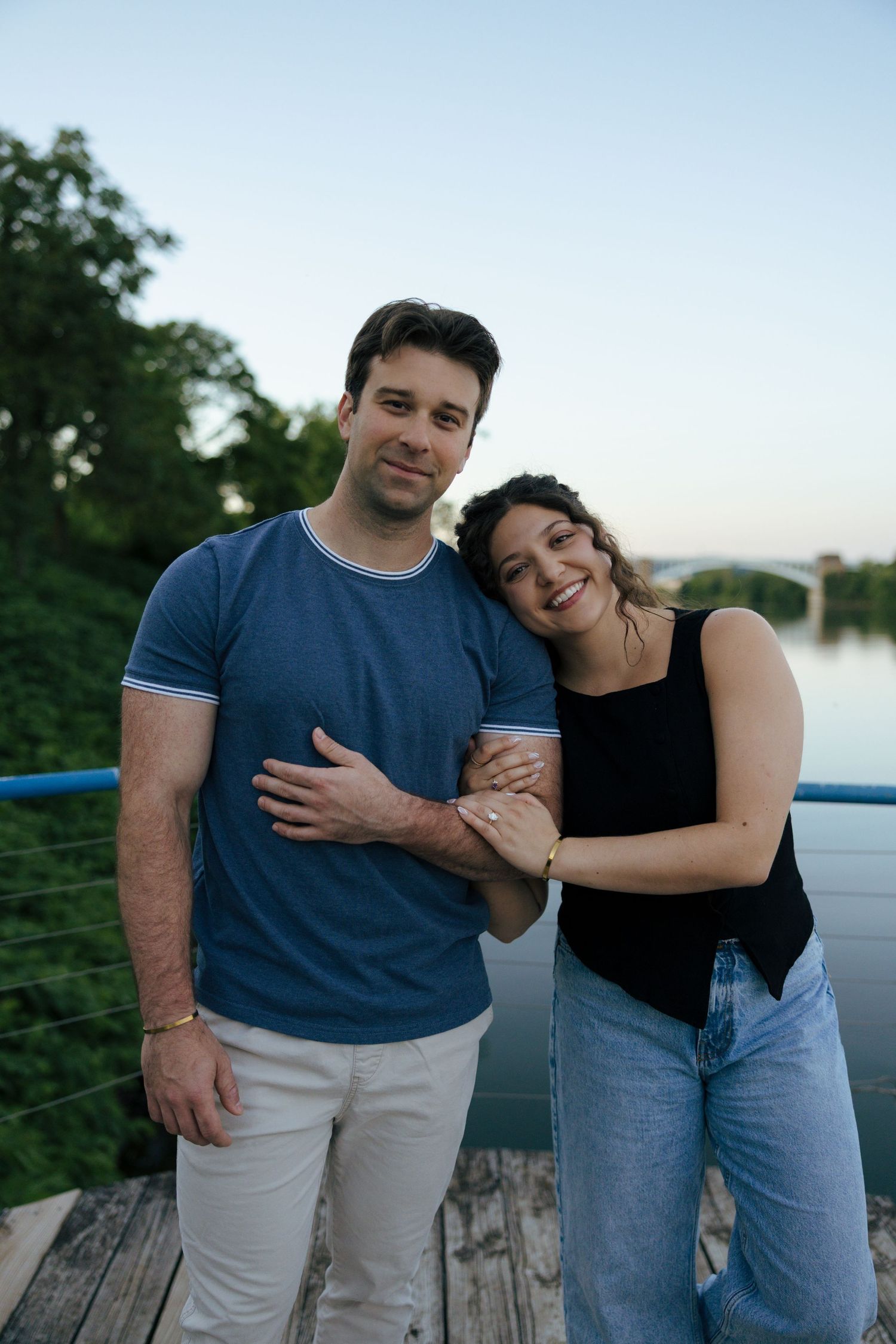 Happy couple embracing on a wooden bridge with a river and trees in the background.