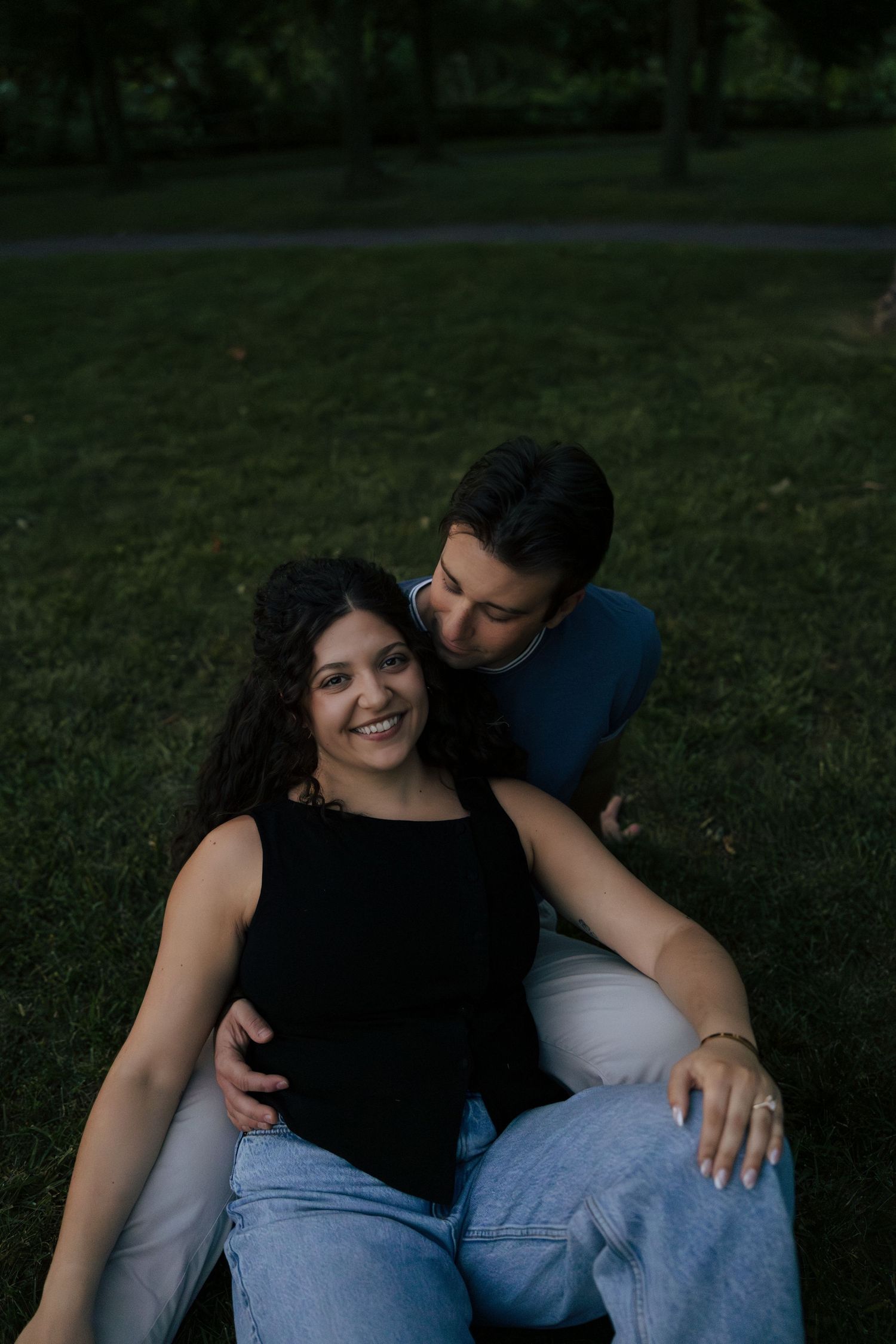 A happy couple sits together on green grass at dusk, the man embracing the smiling woman from behind.