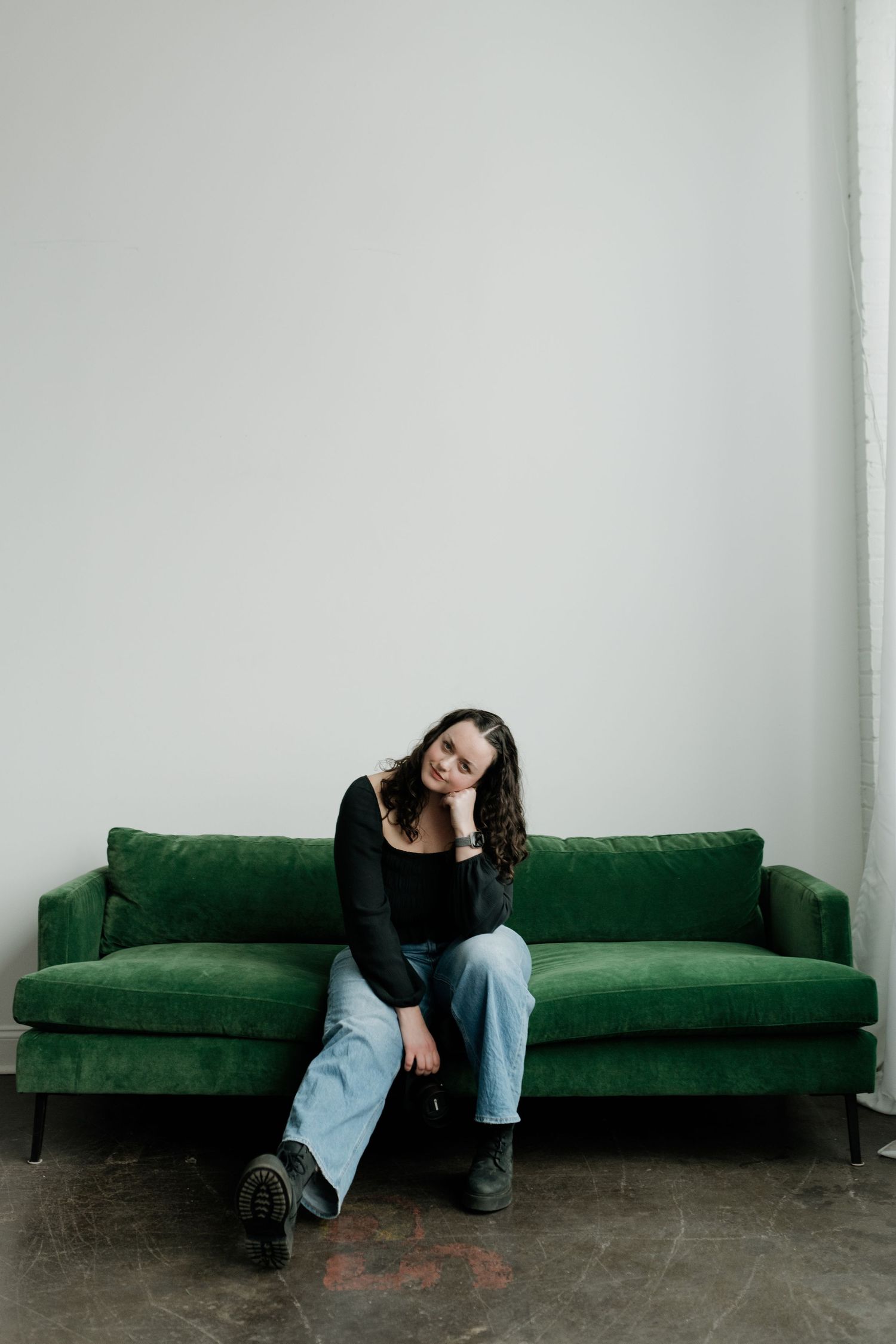 Someone in a black top and light jeans sits casually on a plush emerald green velvet sofa against a white wall.