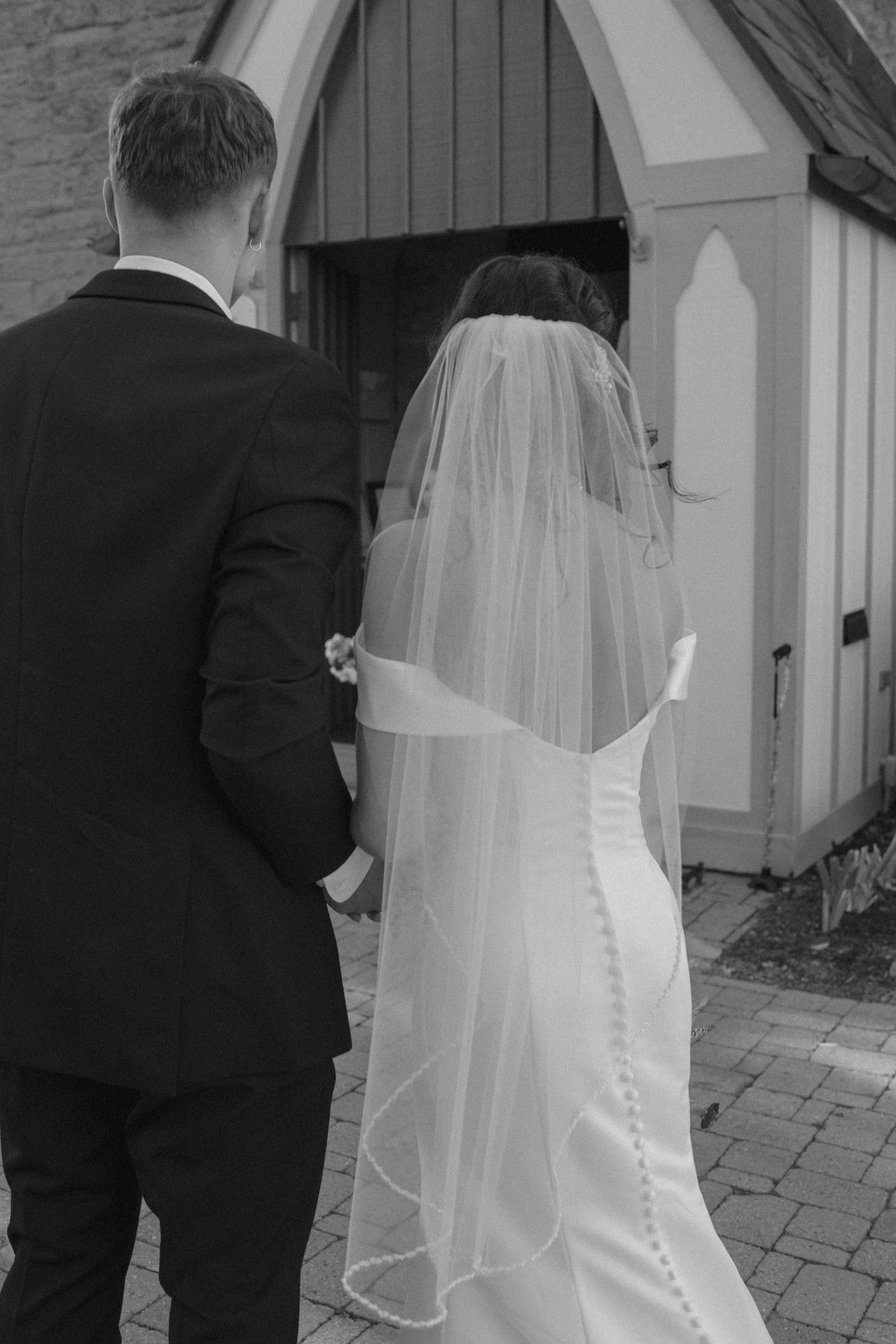 A series of black and white wedding photos showing the back of a bride in a button-back dress with veil and groom in tuxedo.