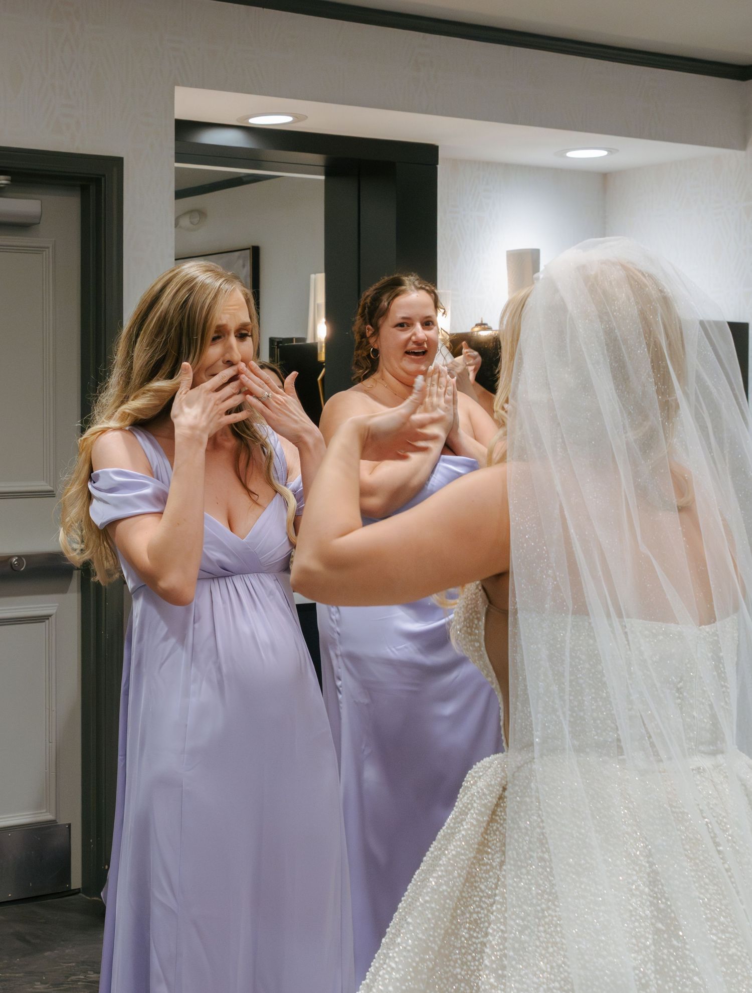 Bridesmaids in lavender dresses help adjust the bride's veil in a bright indoor space.