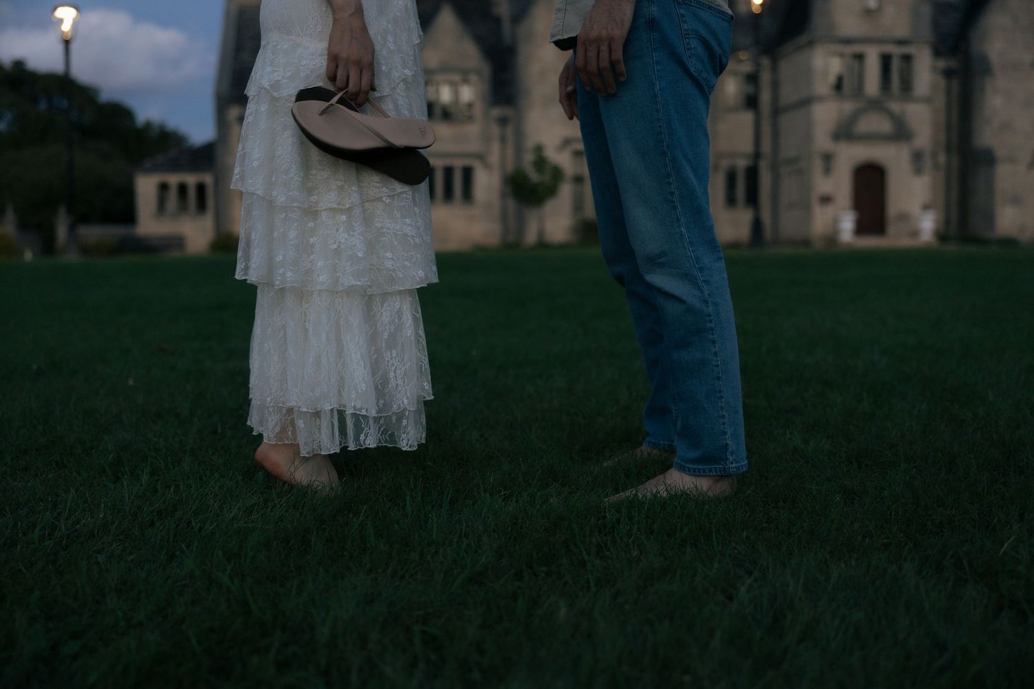 Couple stands on green lawn at dusk before illuminated historic stone castle at twilight.