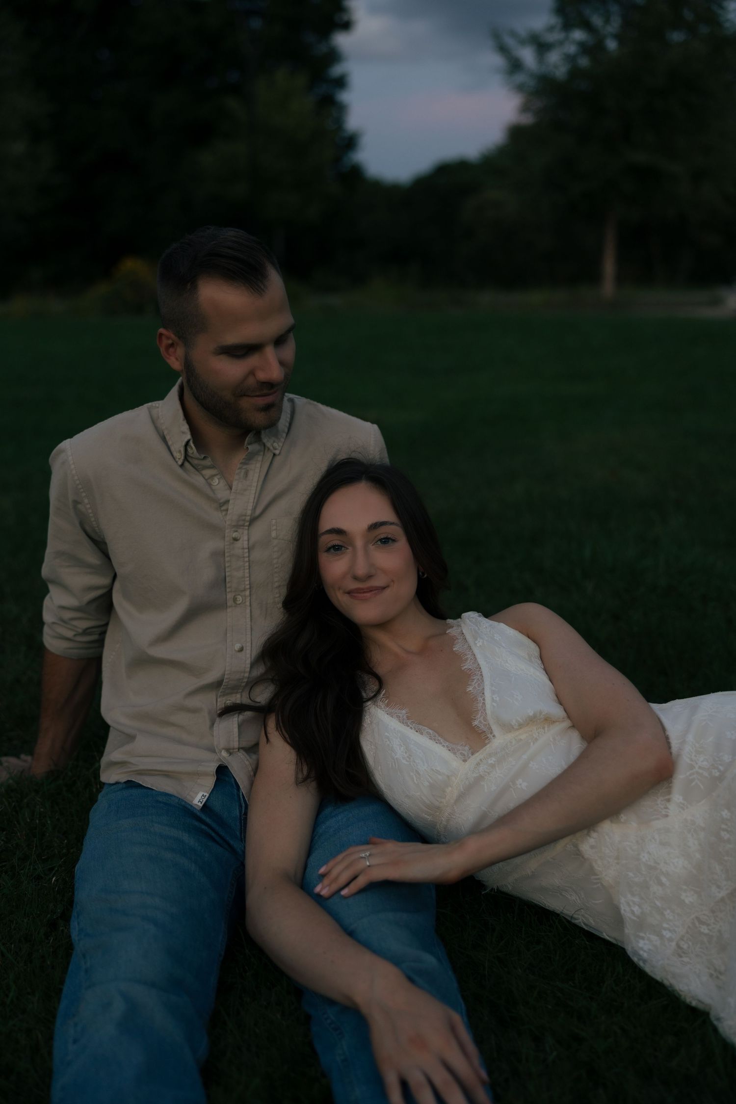 Couple sitting on grass at dusk, woman in white lace dress leaning against man in beige shirt and jeans.